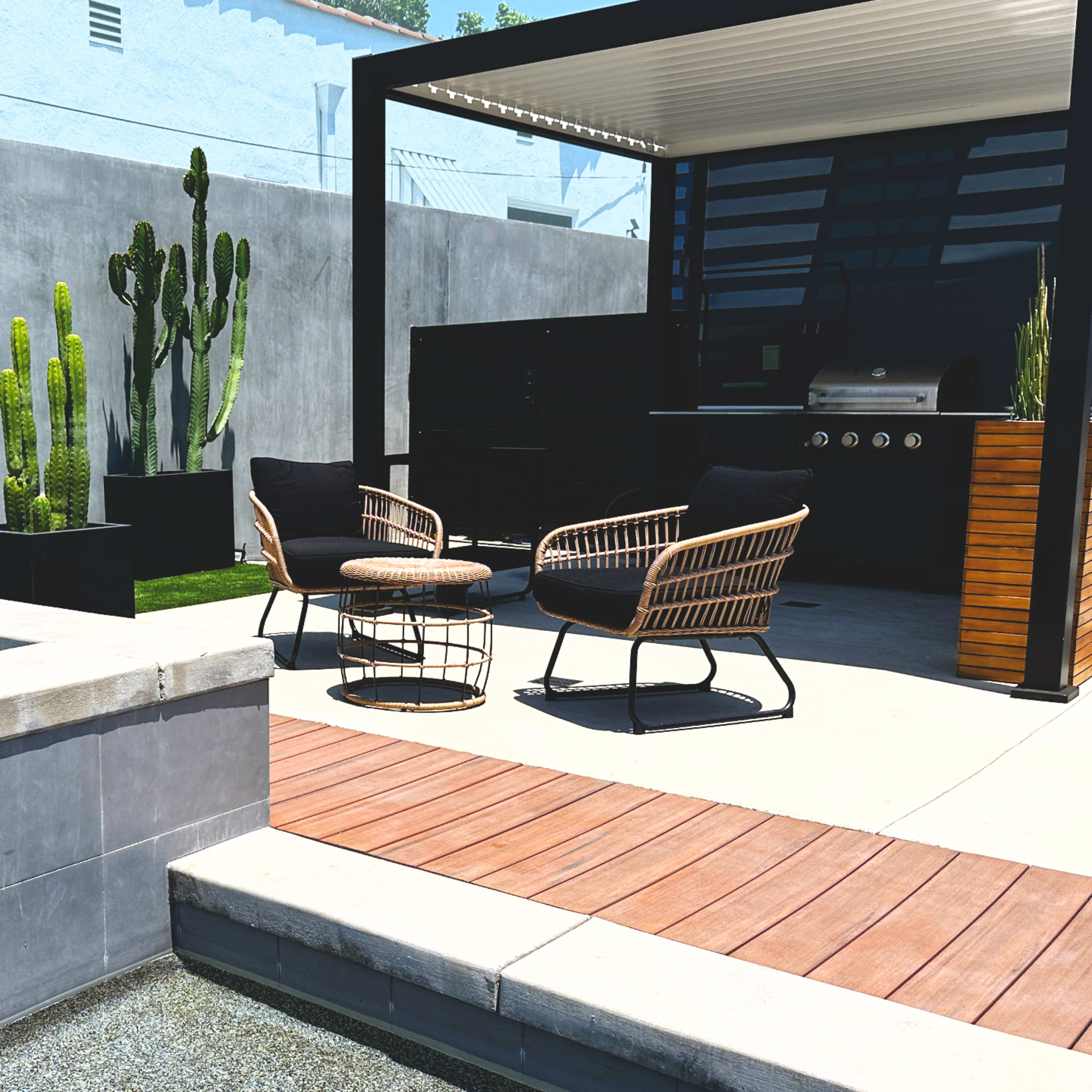 A modern backyard with a seating area beside a small pool, featuring cacti in planters and a barbecue grill under a shaded structure.