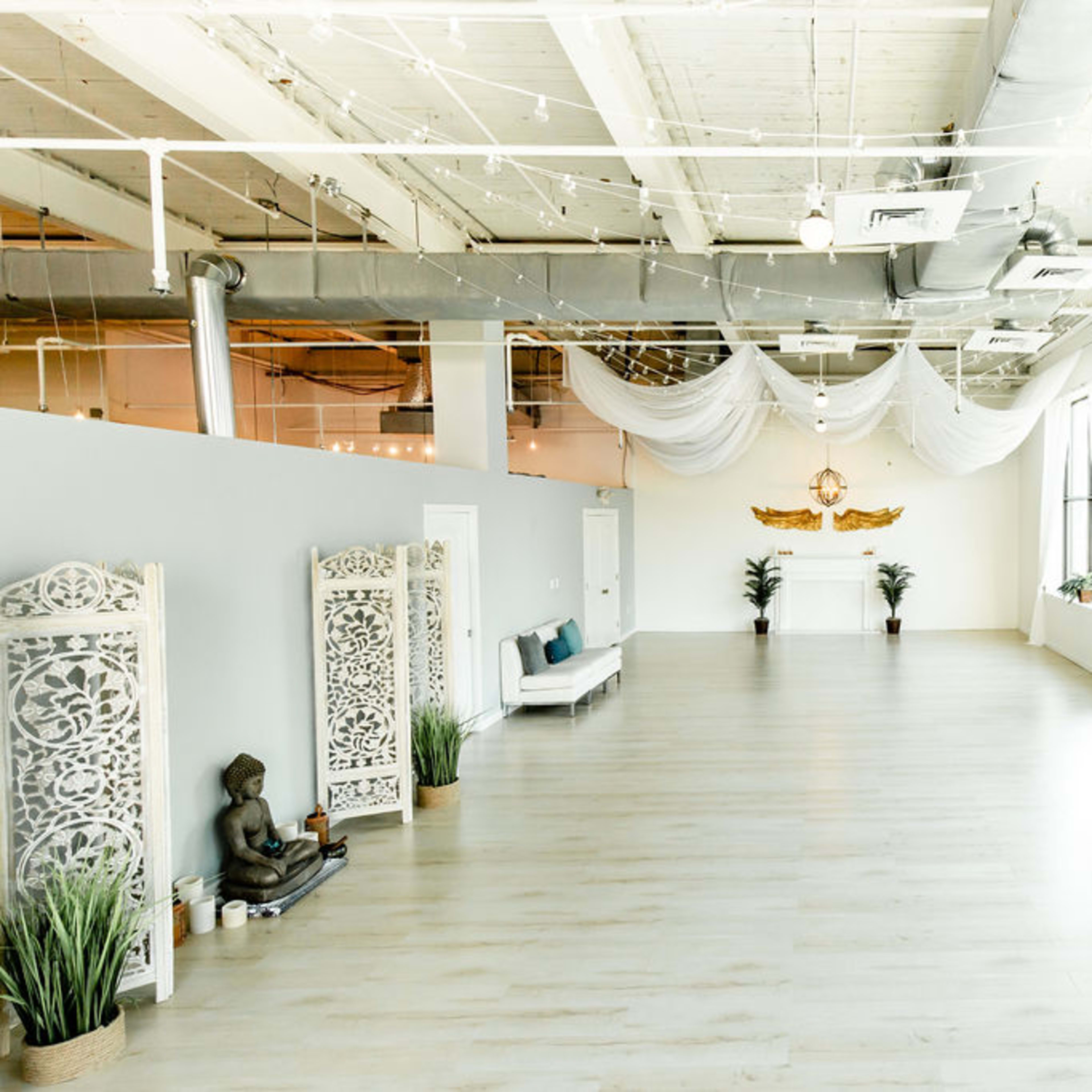 The spacious interior features light-colored wooden flooring, large windows with sheer curtains, and minimal decor including a Buddha statue and ornamental screens.