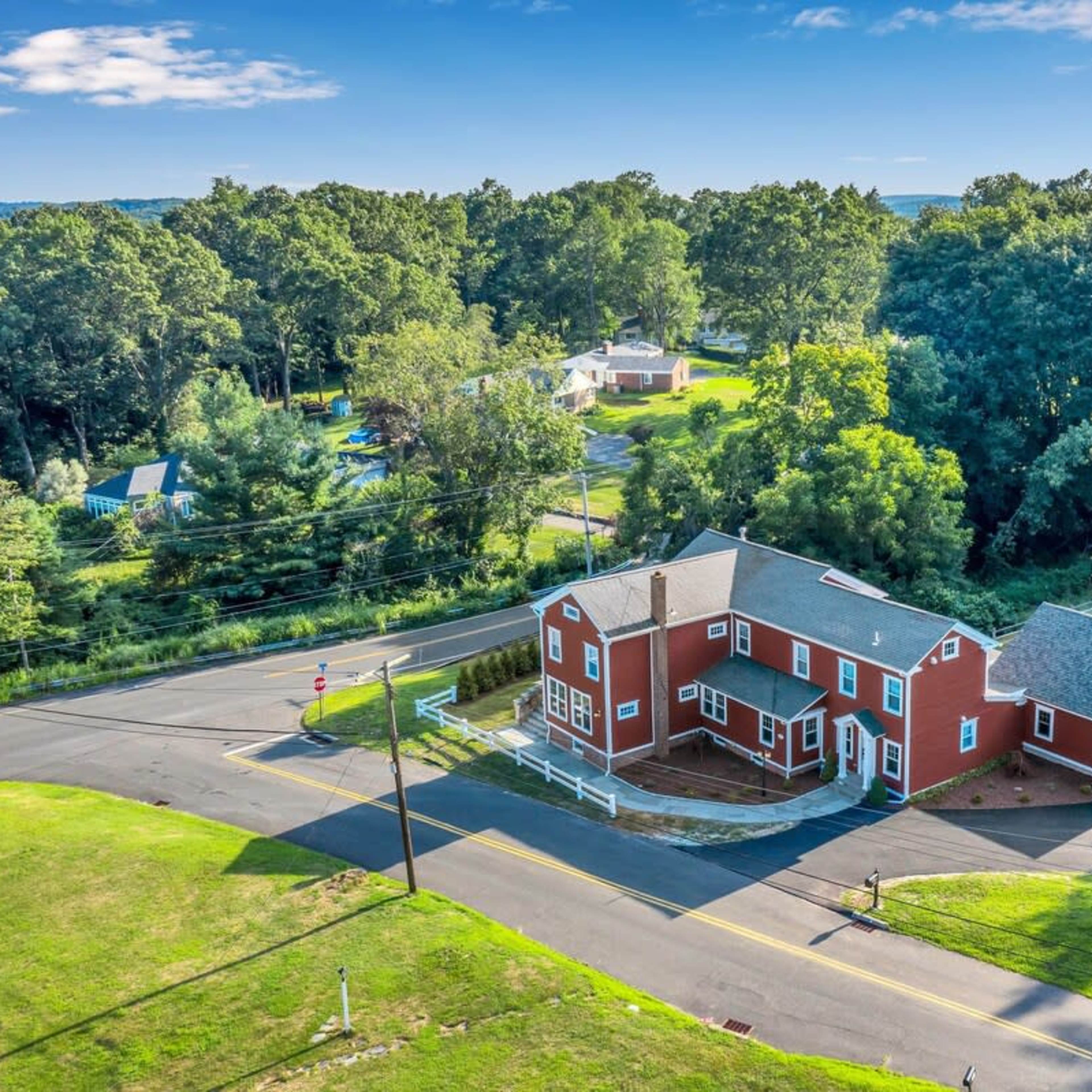 A red two-story house at the intersection of two roads, surrounded by trees and grassy areas.