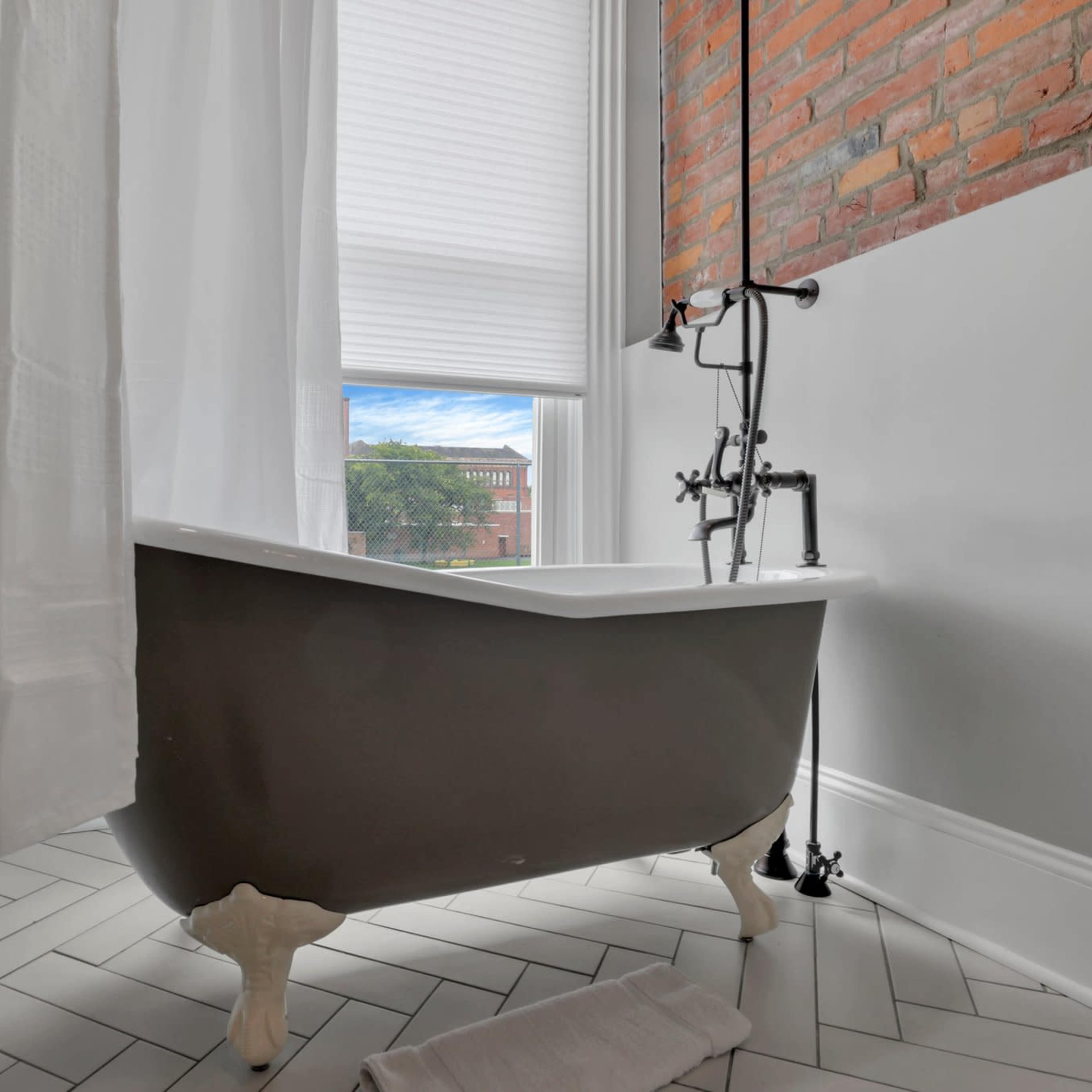 A cast-iron bathtub with claw feet is positioned beside a window, partially obscured by white curtains, in a room with a brick and painted wall.