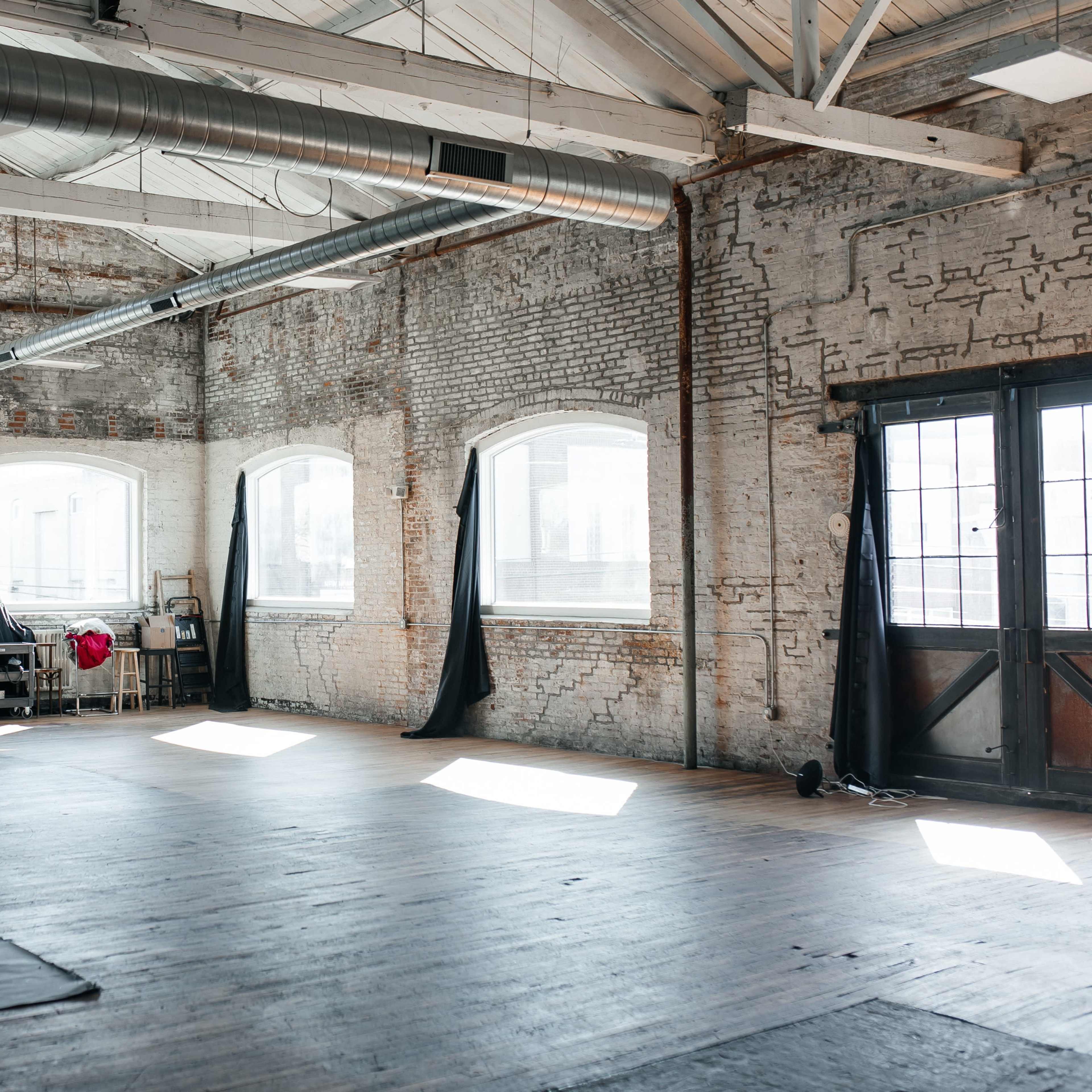 An empty studio space with exposed brick walls, large windows, and wooden floors, featuring a backdrop and lighting equipment set up in one corner.