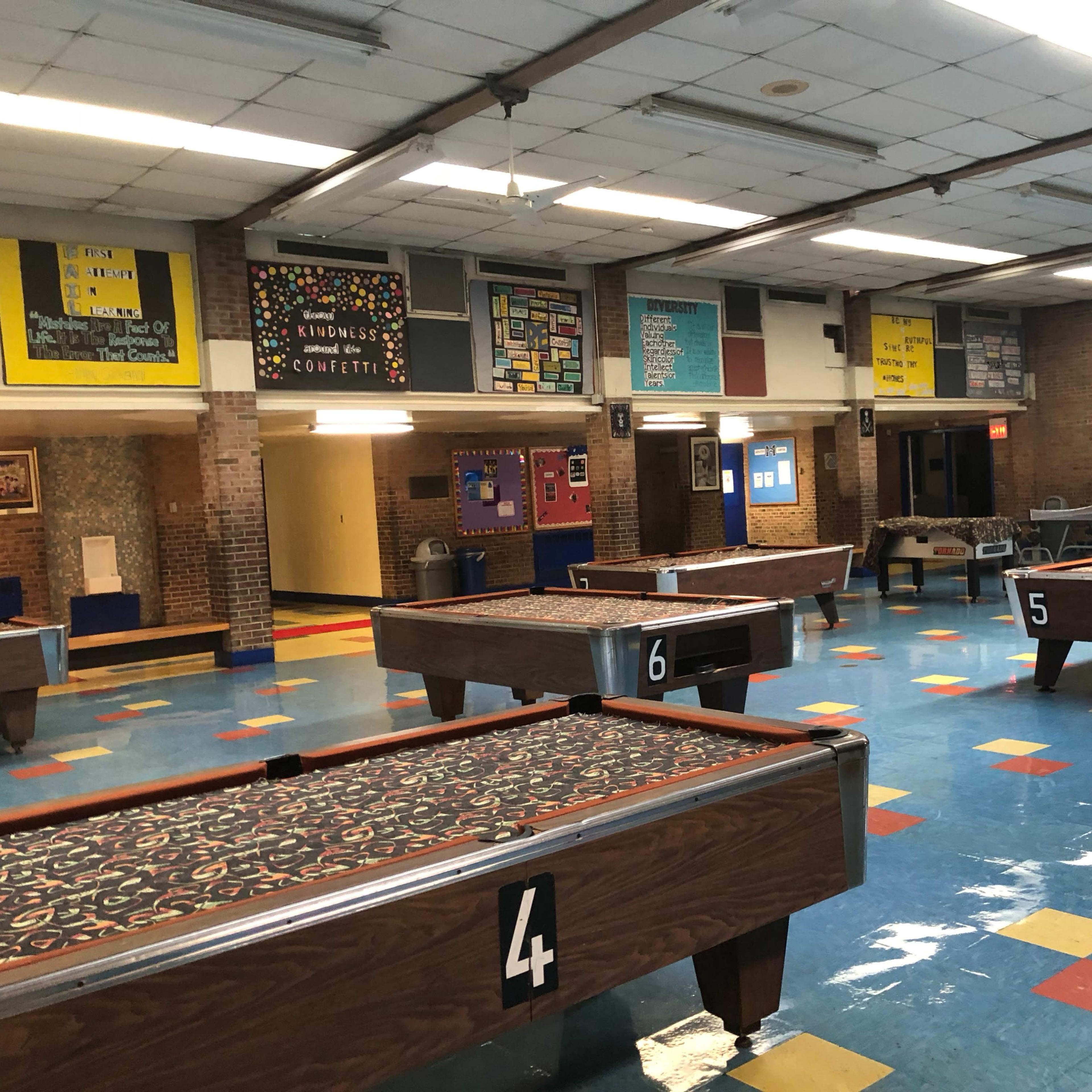 An empty gymnasium with several pool tables labeled with numbers, colorful floor tiles, and bulletin boards on the walls.