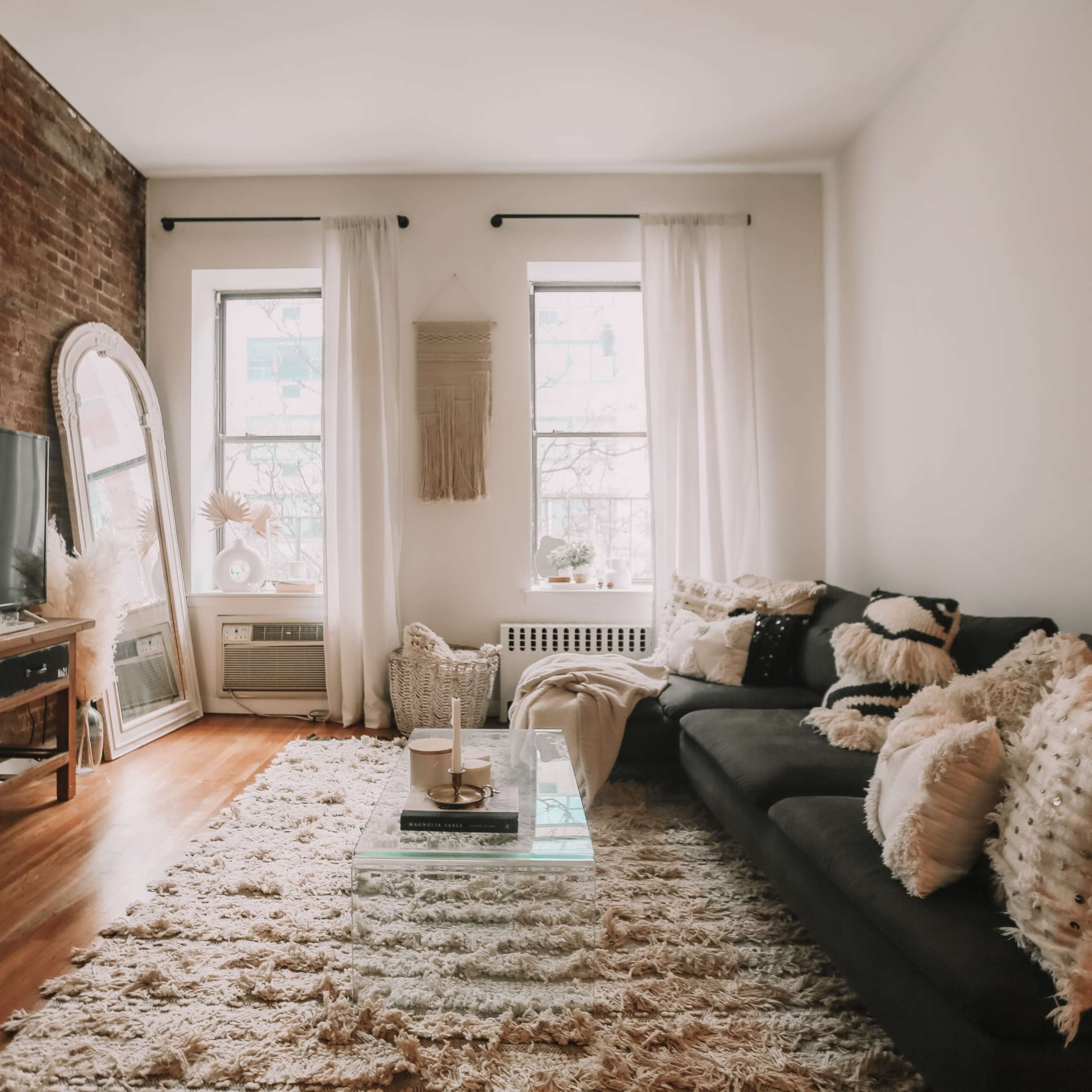 The living room features a dark sofa adorned with patterned pillows, a glass coffee table on a textured rug, and exposed brick walls with large windows.