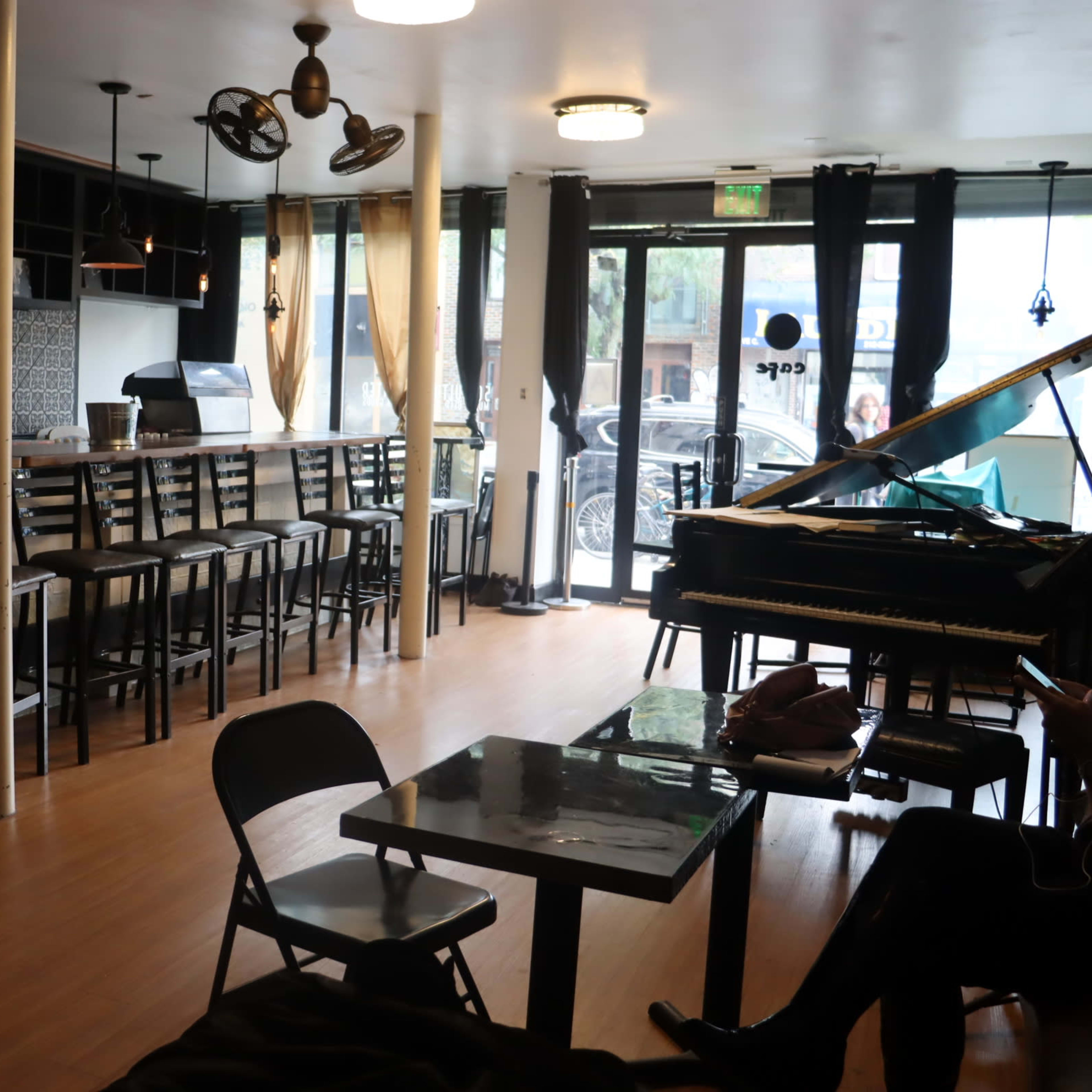 A spacious café interior with a long counter, bar stools, a piano, and large windows allowing natural light to enter.