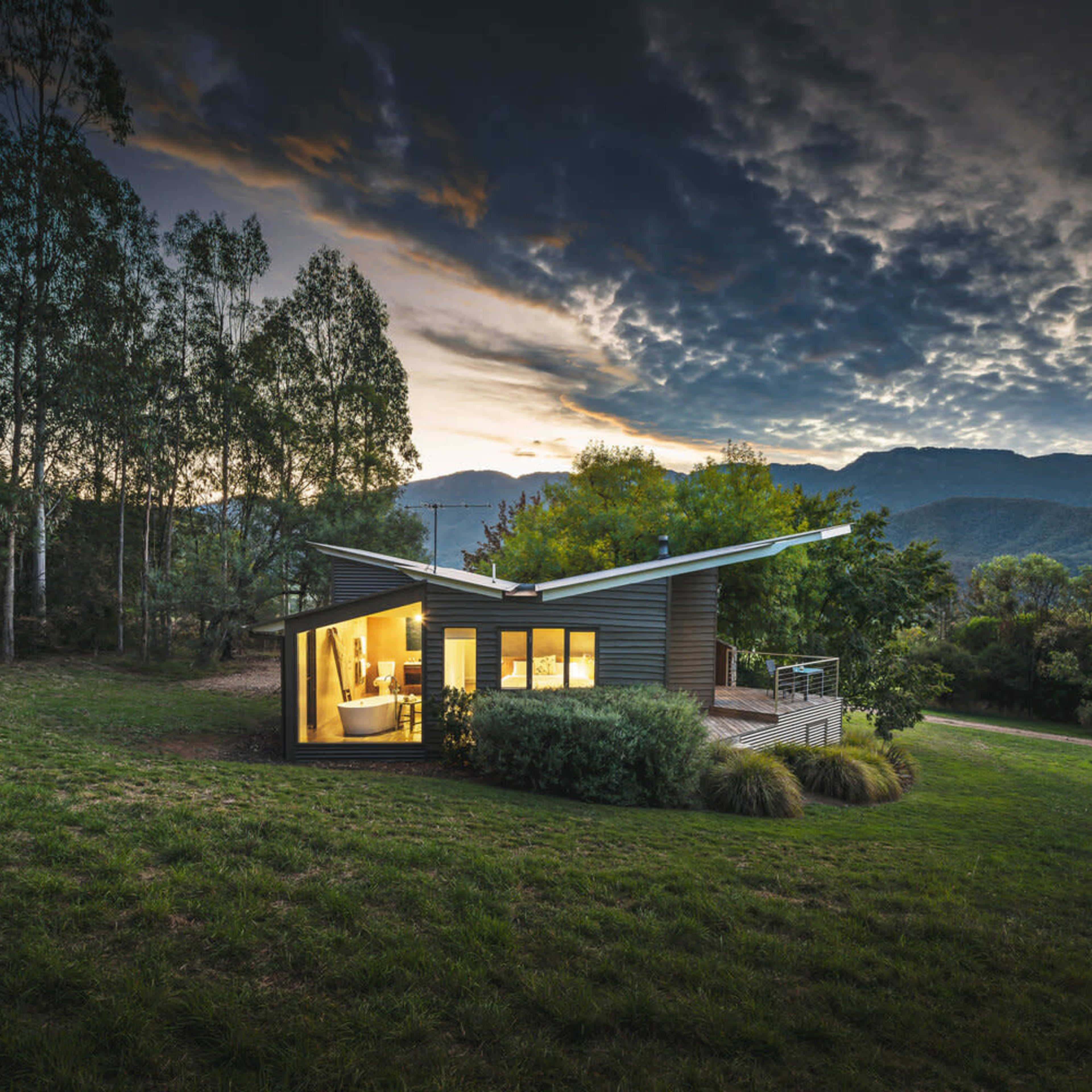 A modern, angular house with large windows is situated in a grassy area surrounded by trees, with mountains in the background under a cloudy sky.