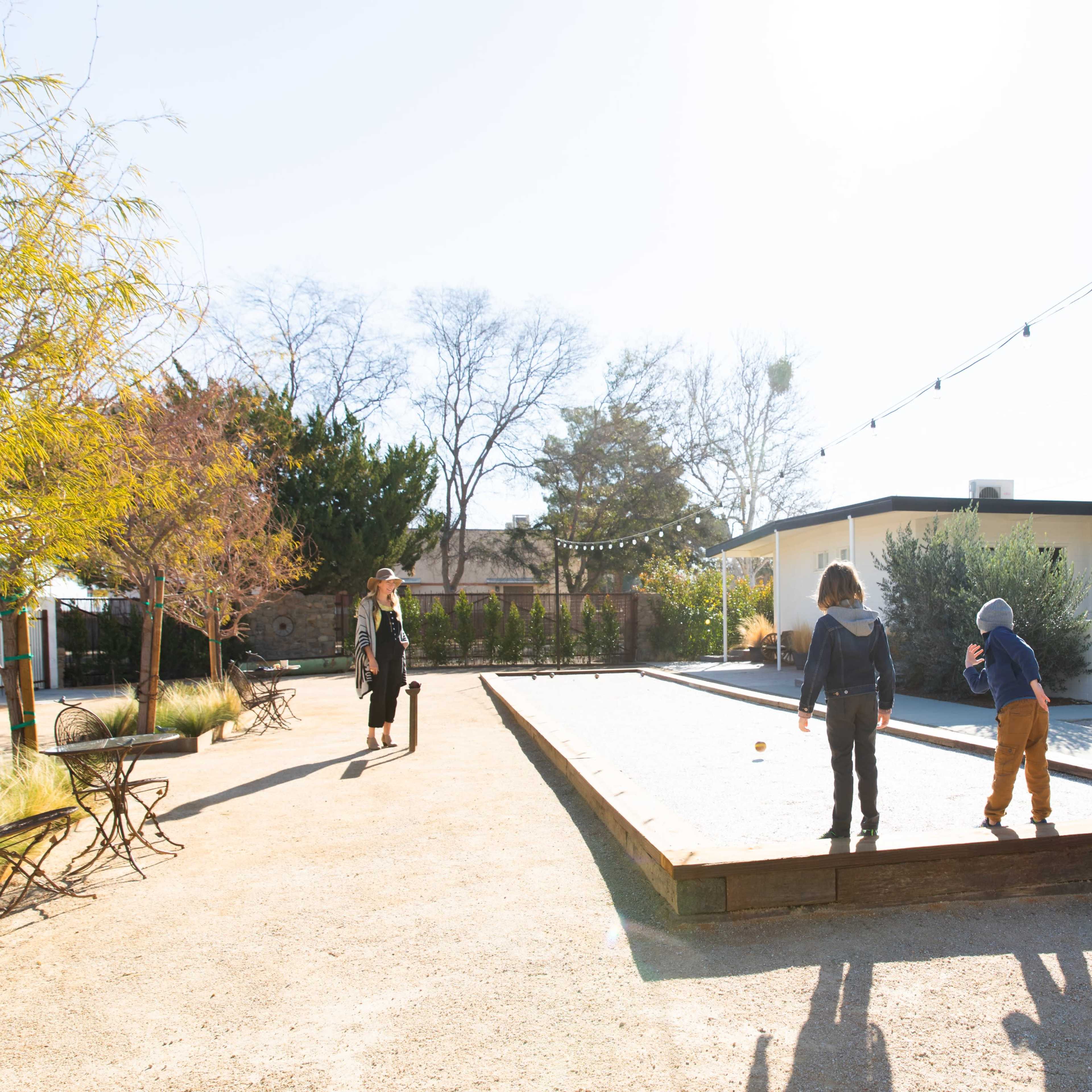 A man and two children are playing a game on a gravel court while a woman walks in the background.