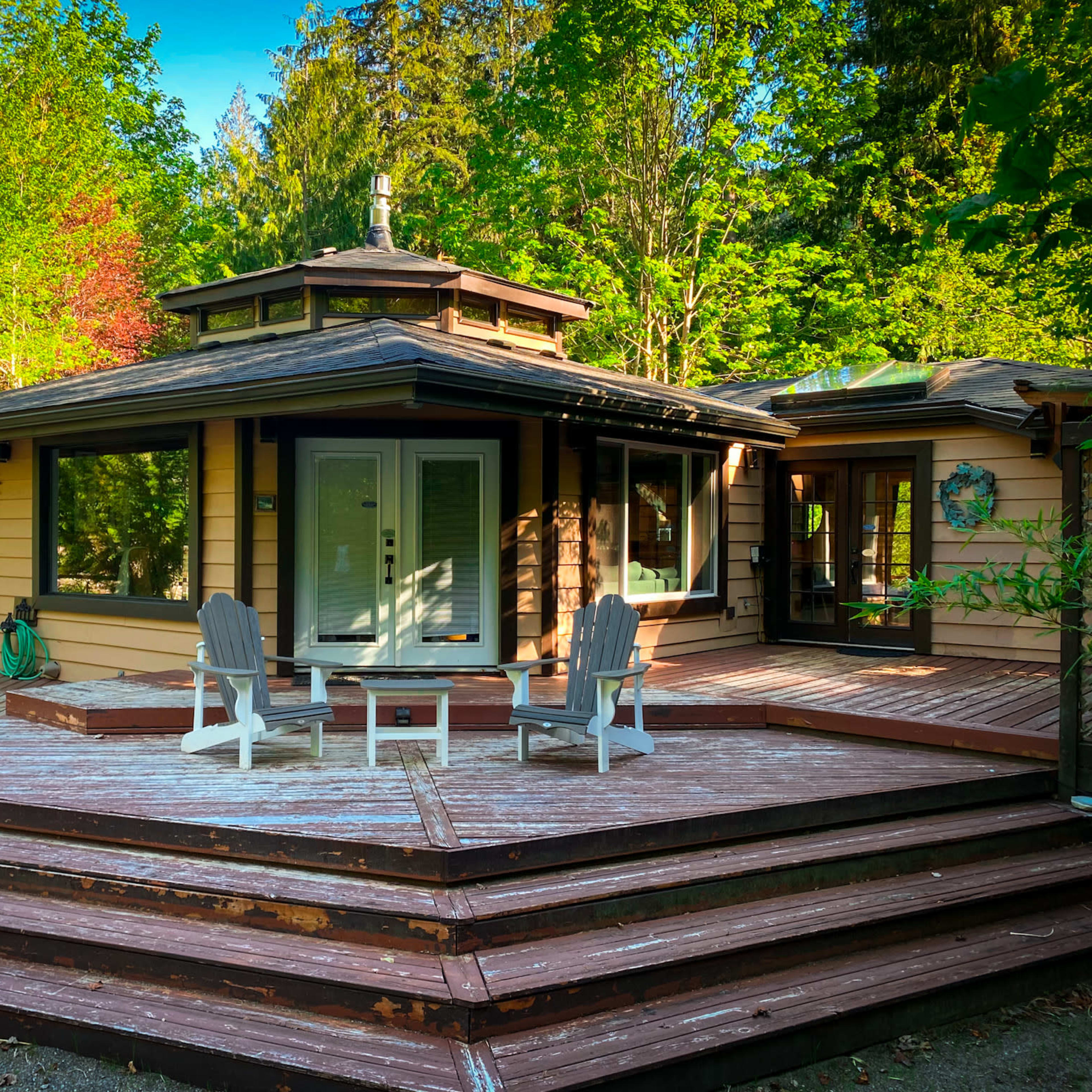 A modern, single-story house surrounded by trees, with a wooden deck featuring a seating area in front.