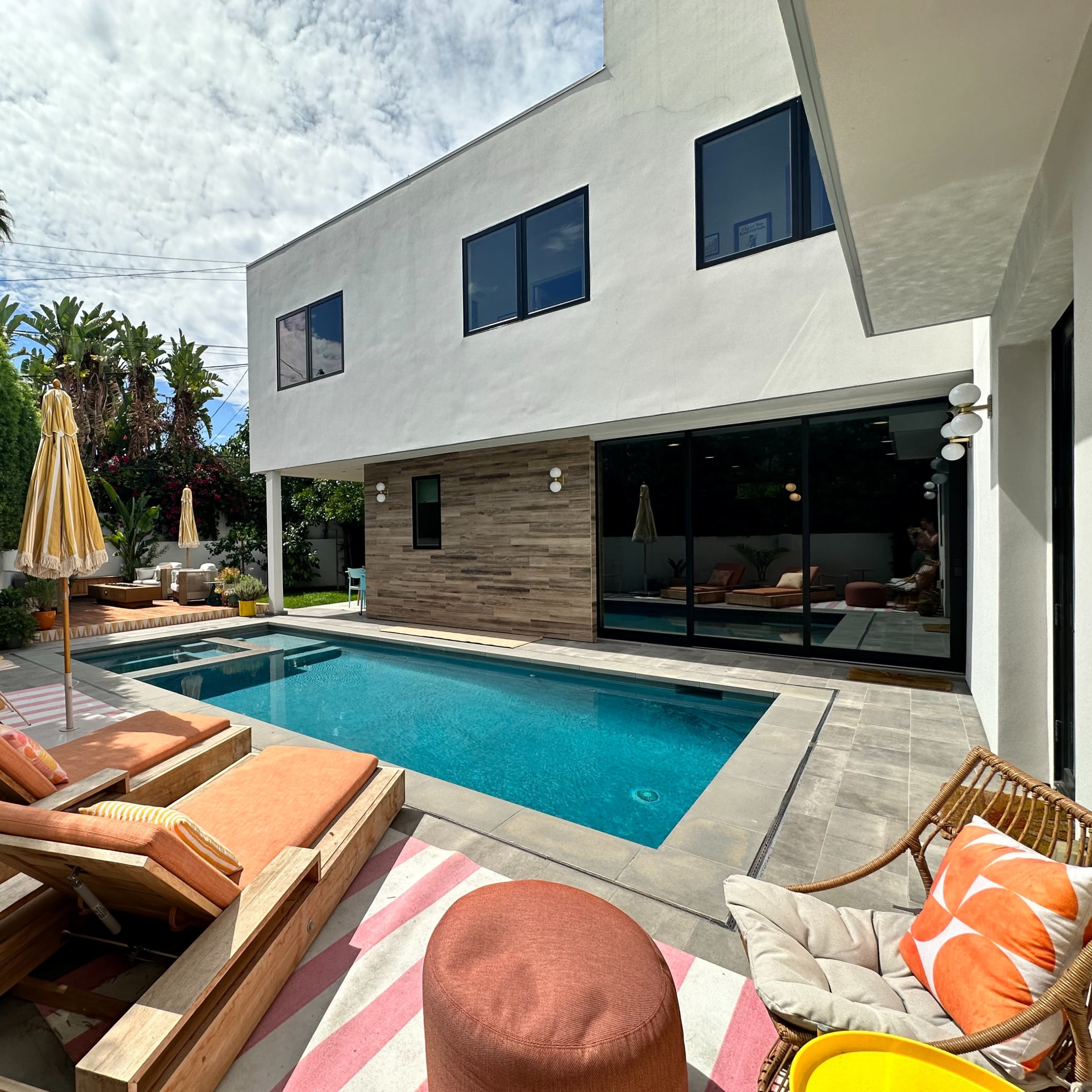 A modern backyard features a swimming pool, lounge chairs, and a patio area with seating, surrounded by lush greenery.