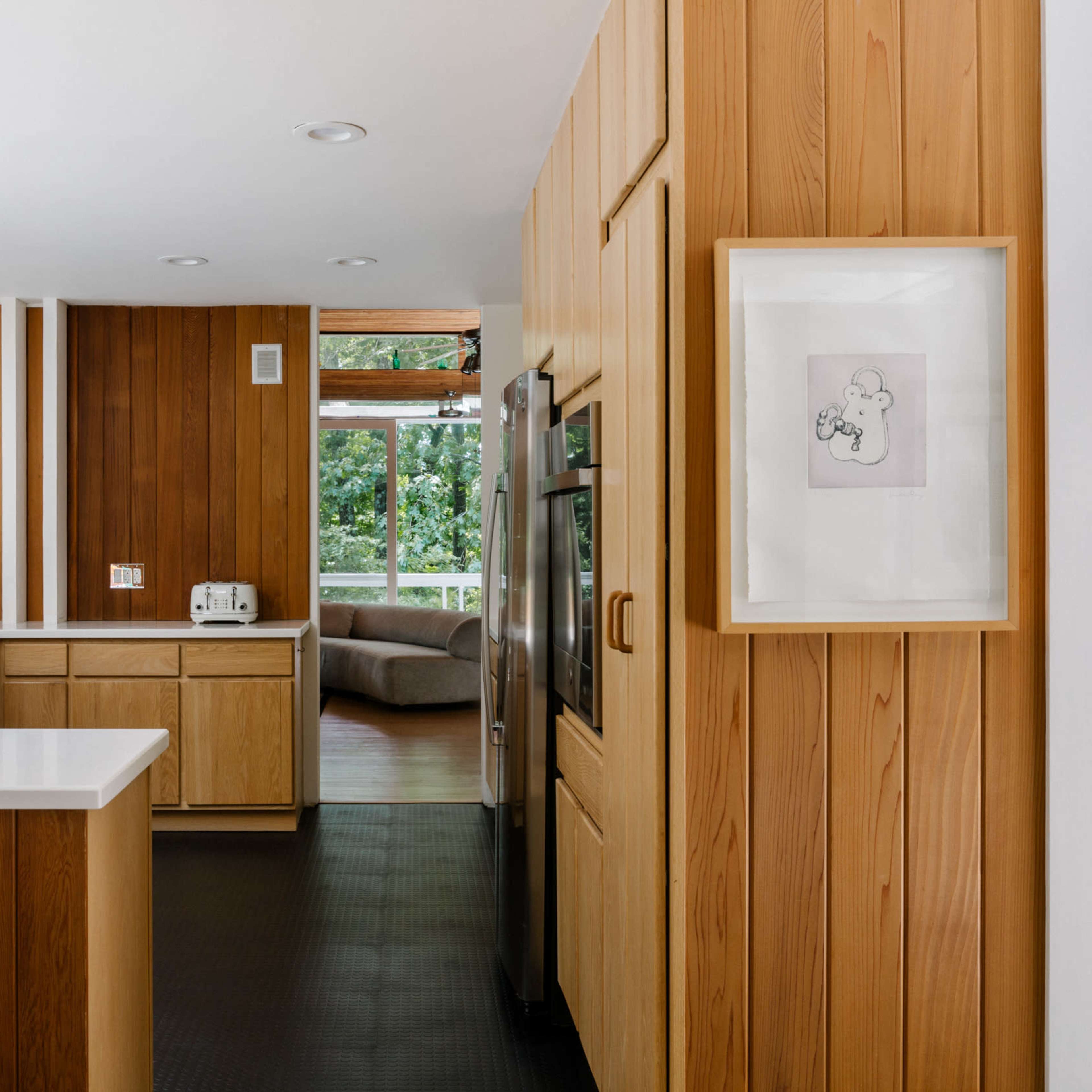 The kitchen features wooden paneling, a white countertop, and stainless steel appliances, with a view of a living area through a large window.