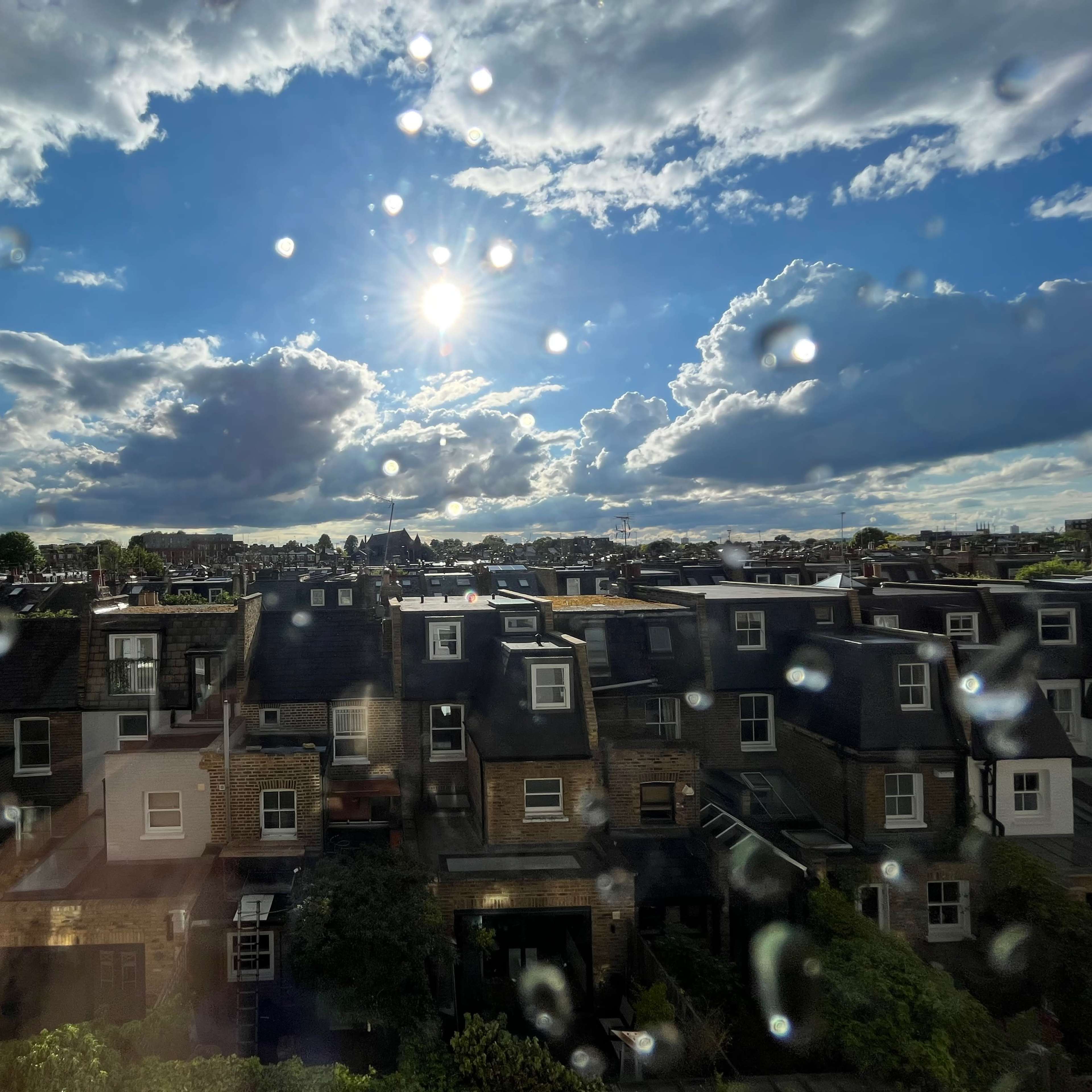 A view of rooftops with a bright sun and scattered clouds against a blue sky, with water droplets on the window.