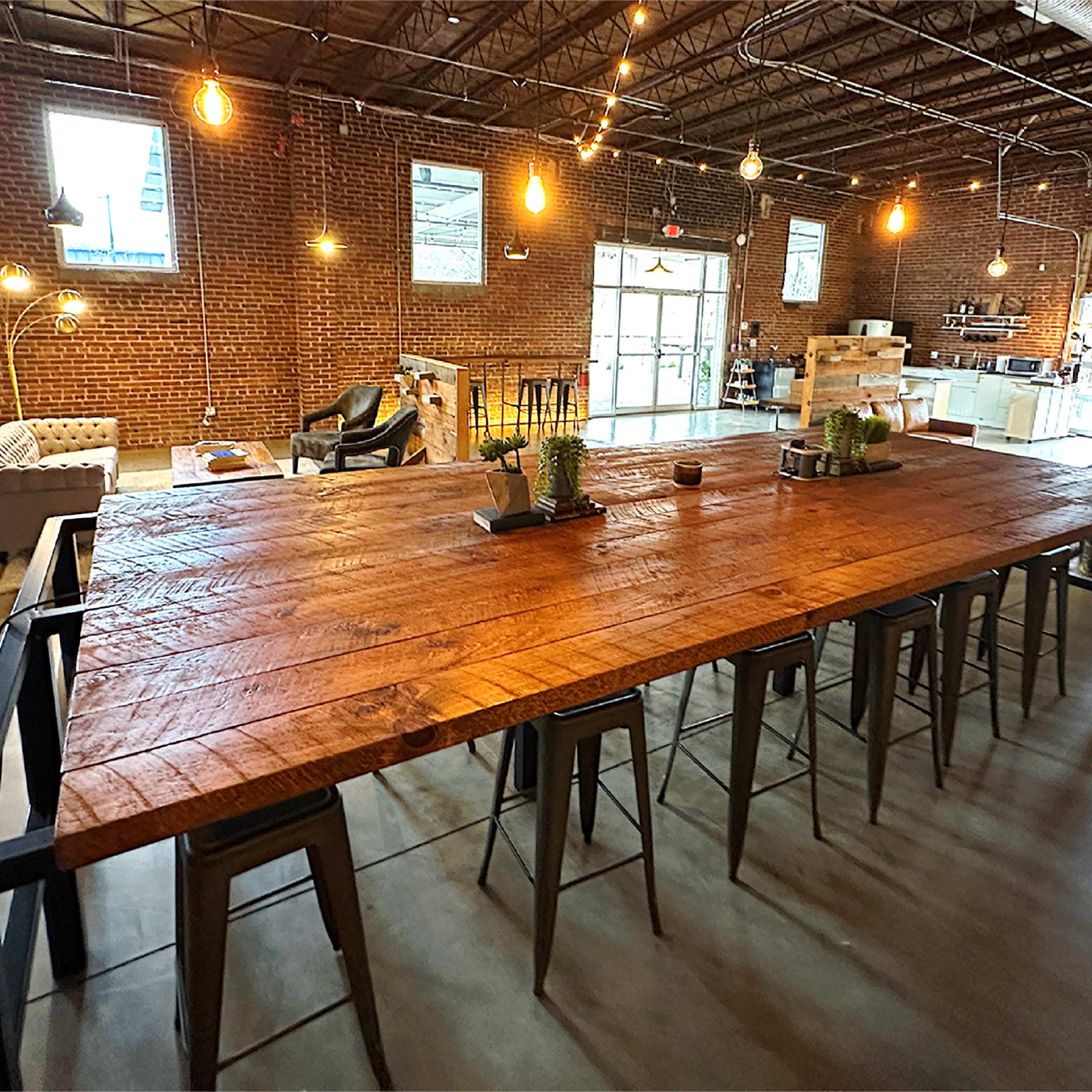 A large wooden table with metal stools is positioned in a spacious, industrial-style room with exposed brick walls and hanging light fixtures.