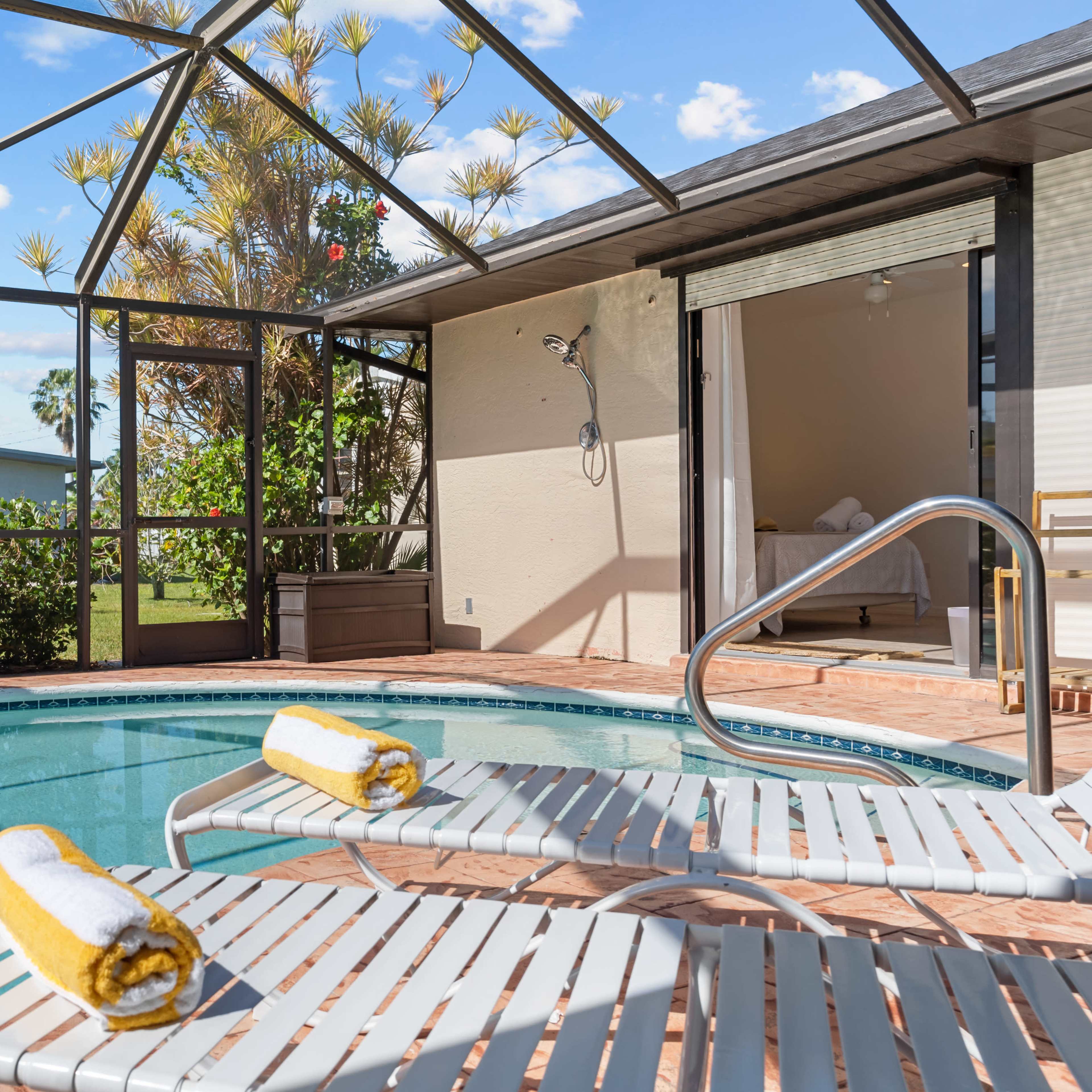 A swimming pool with two yellow towels on lounge chairs is situated within a screened enclosure, with a view of lush greenery and a house in the background.