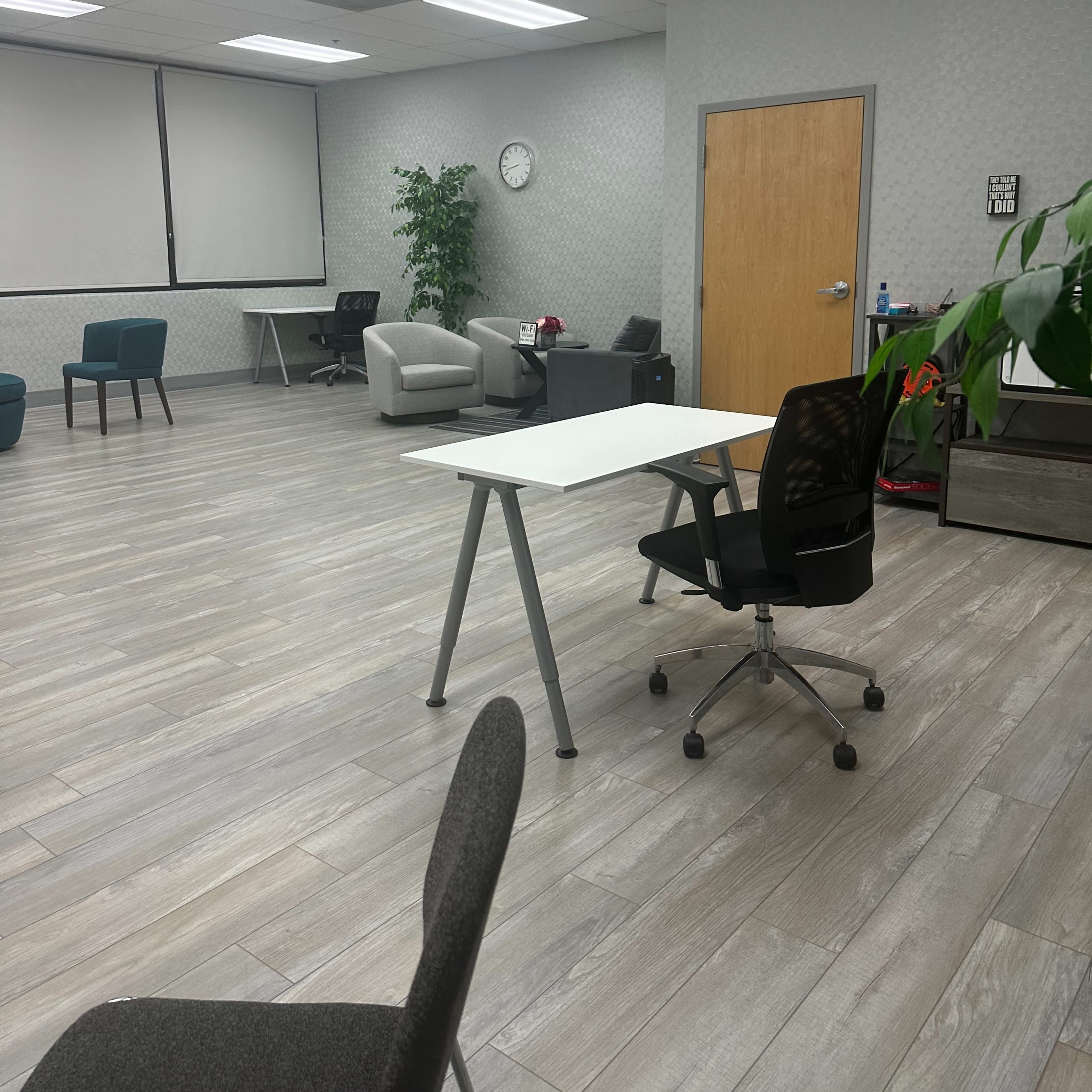 A modern office space features a white table and chairs, with a combination of seating areas and greenery along the walls.