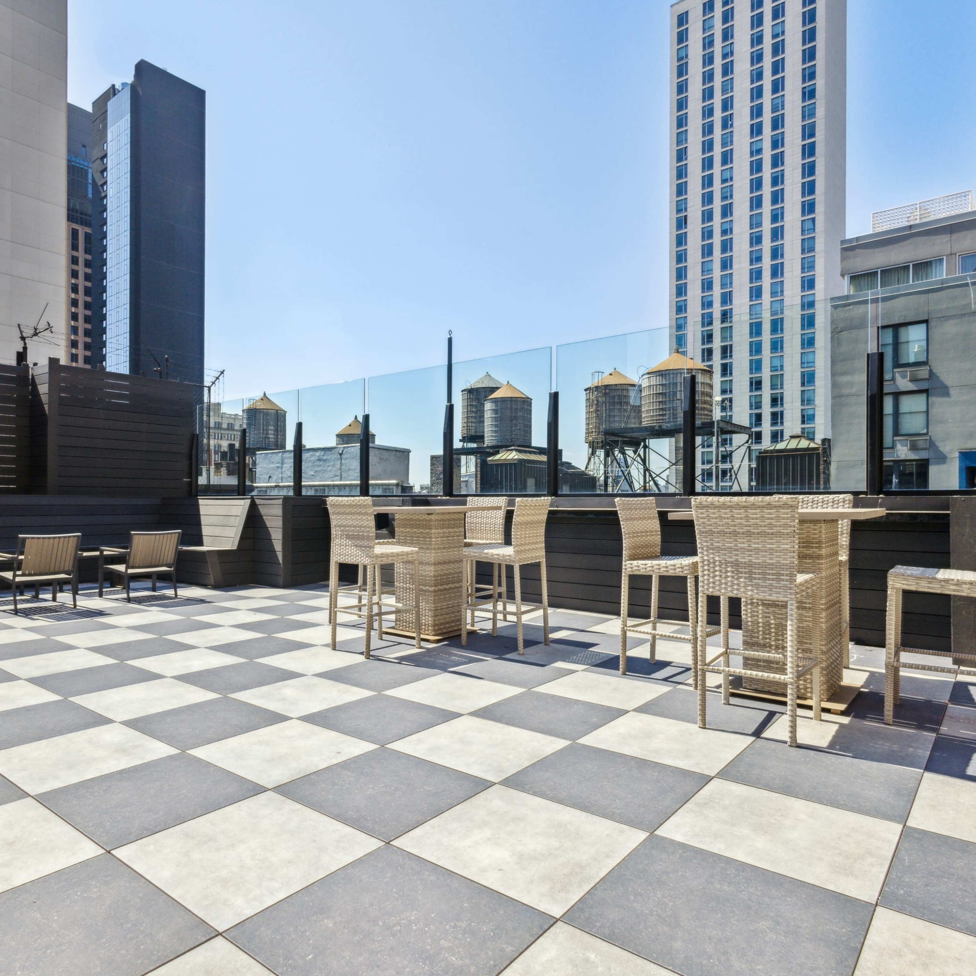 A rooftop terrace featuring a checkered tile floor, tall seating areas, and a view of surrounding buildings and water towers.