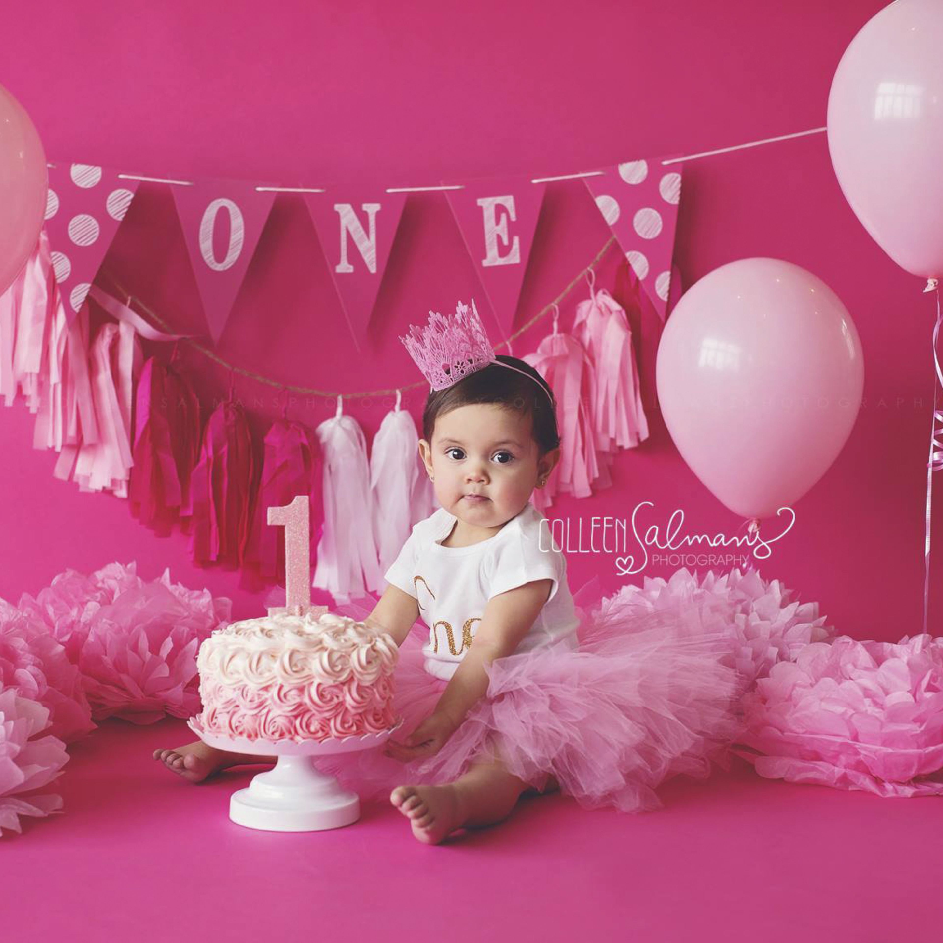 A baby girl sits in front of a pink backdrop adorned with balloons, a decorative banner, and a cake topped with the number one.