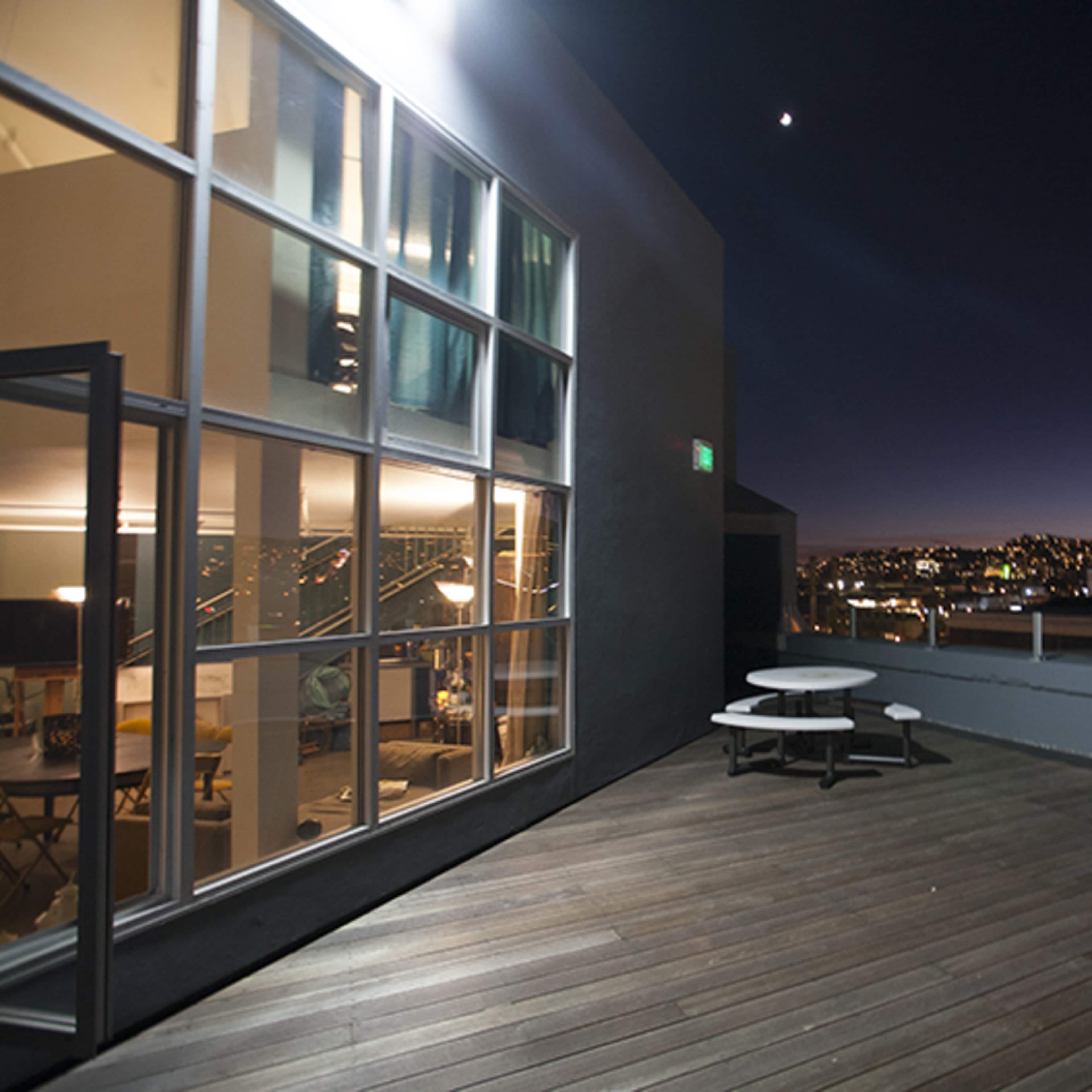A rooftop patio with a circular table and chairs overlooks a city skyline at night, illuminated by distant lights.