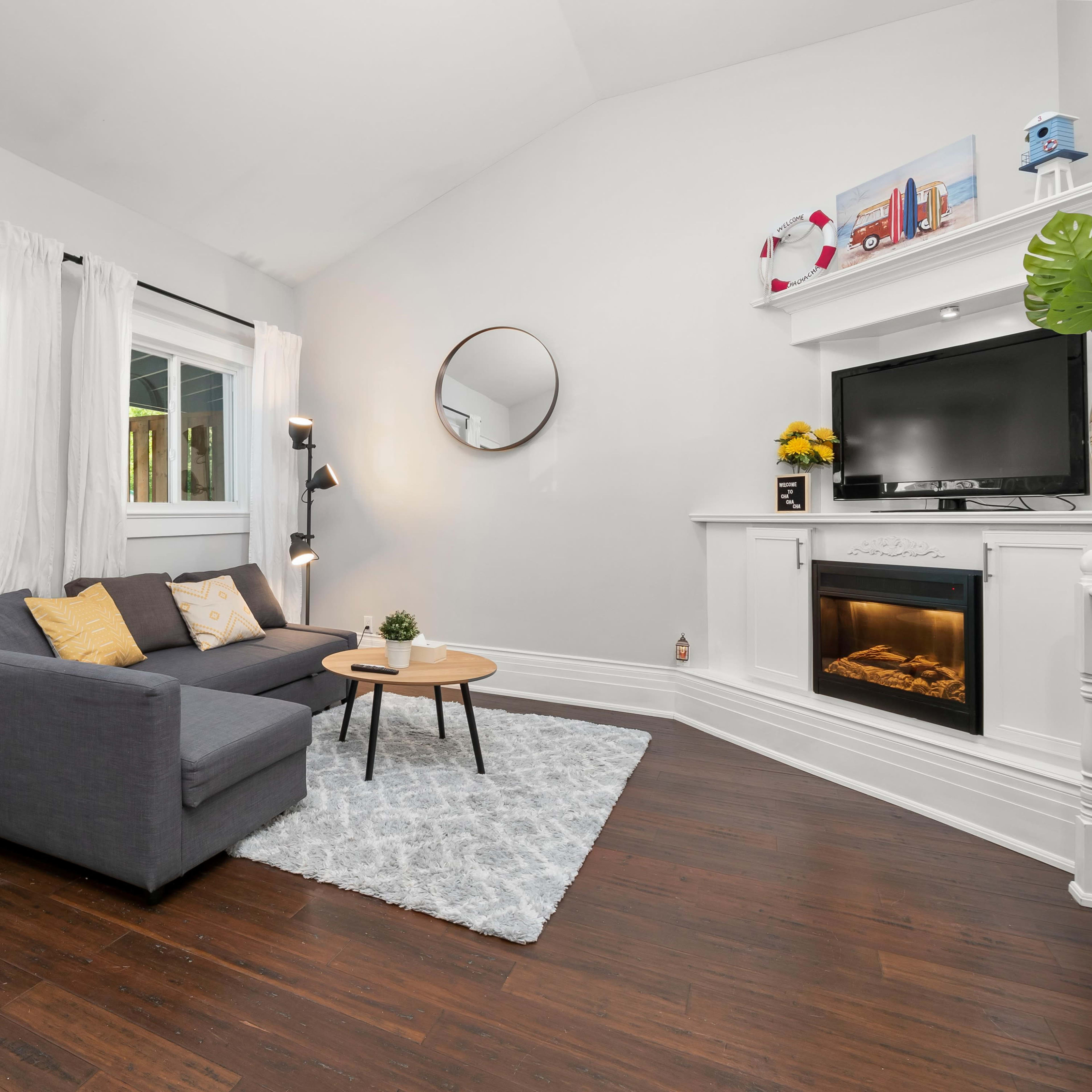 A modern living room with a gray couch, a round mirror, a television mounted on a white fireplace, and large windows allowing natural light to enter.