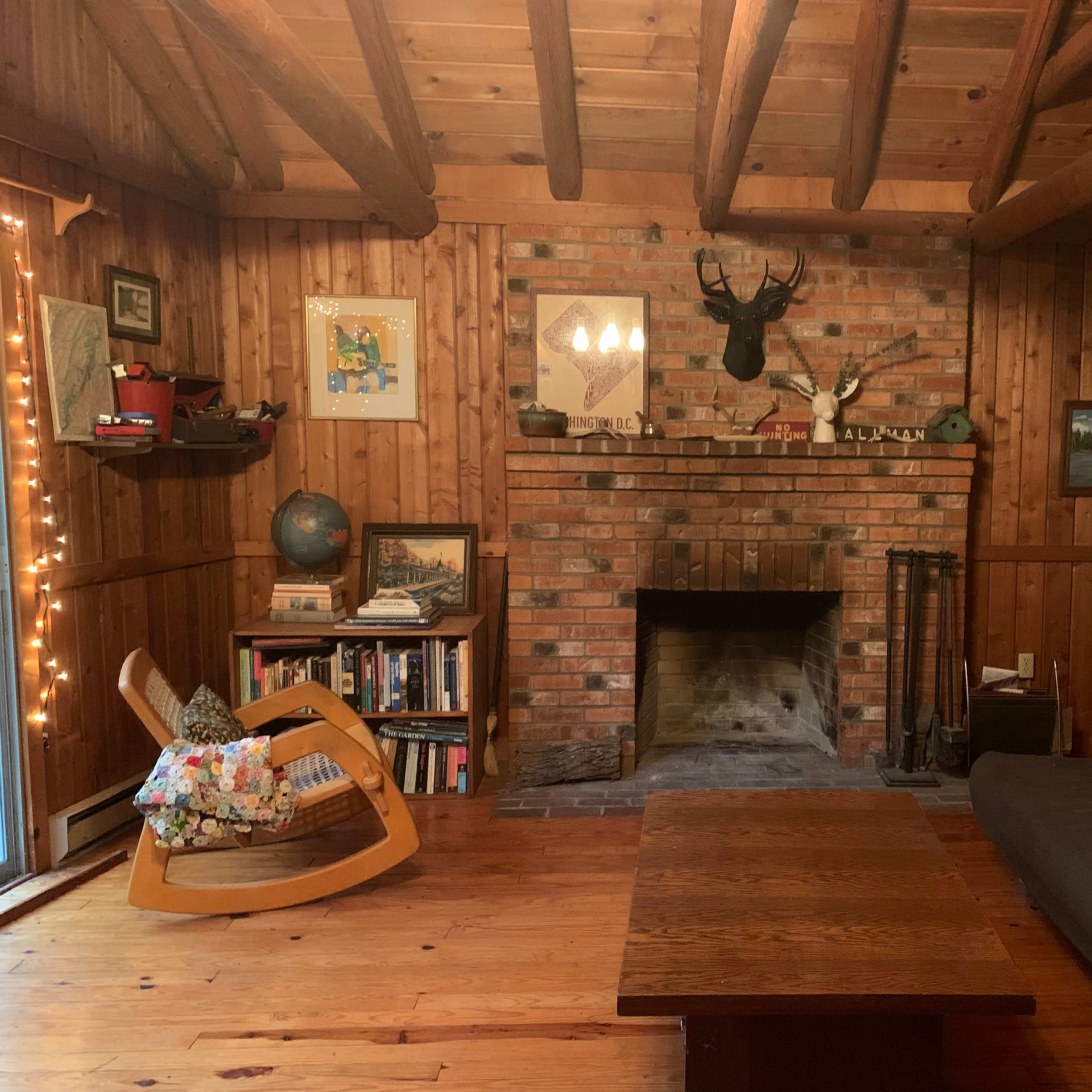 The living room features a brick fireplace, wooden beams, a rocking chair, and a wooden table surrounded by bookshelves and wall art.