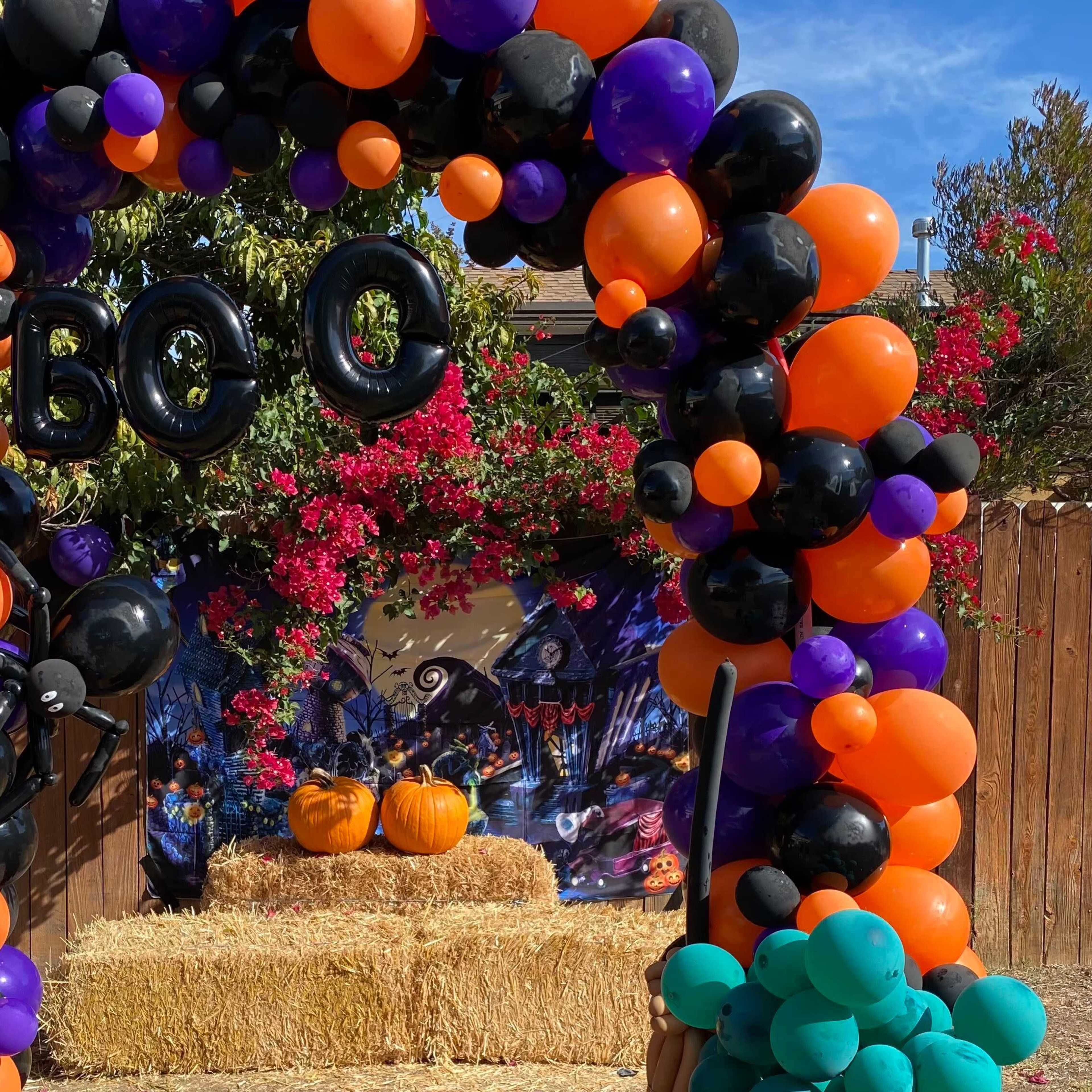 A decorative arch made of orange, black, and purple balloons frames a display of pumpkins on hay bales, set against a backdrop of flowers and Halloween-themed decorations.
