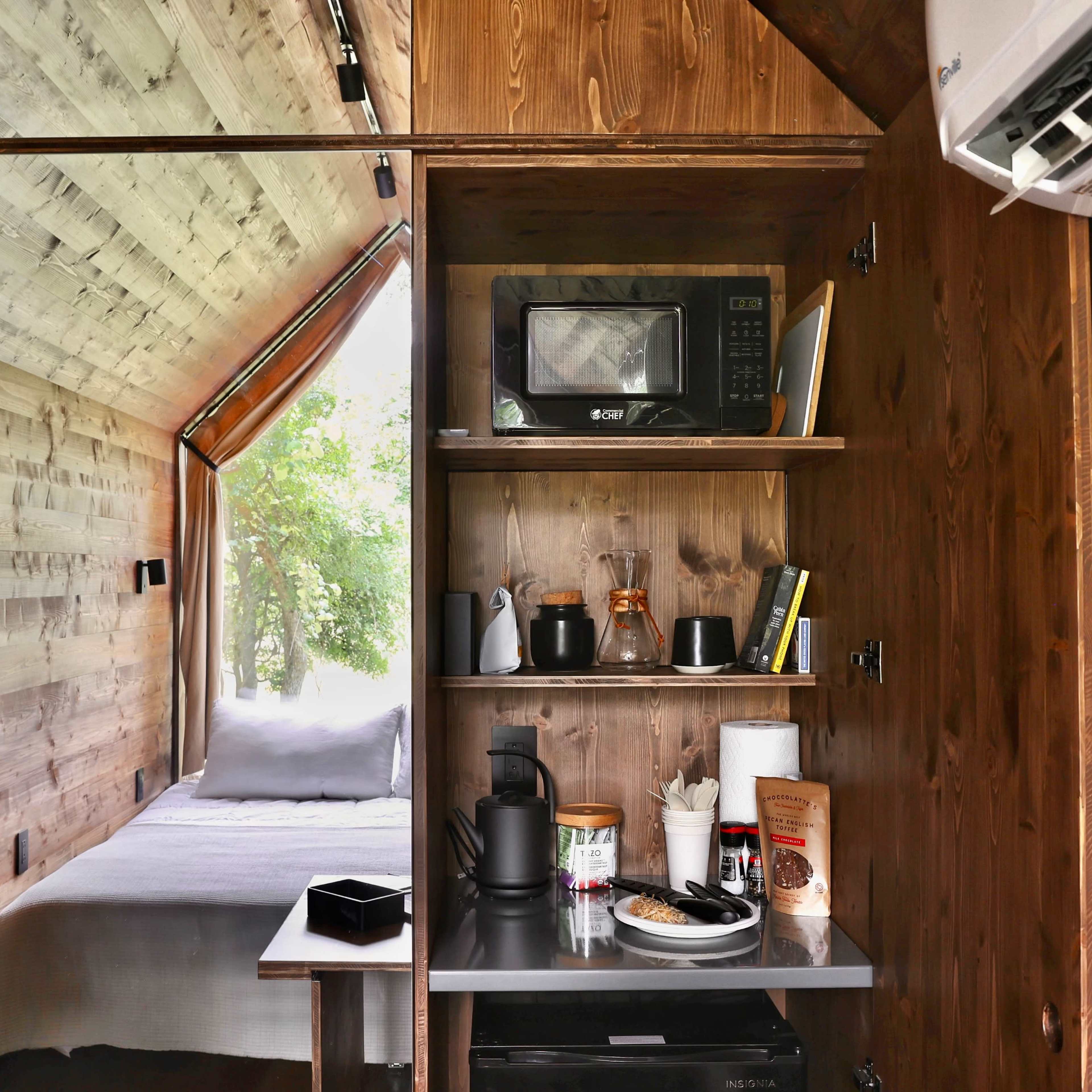A compact wooden interior featuring a kitchenette with a microwave, coffee maker, and food items, alongside a small bedroom area with a bed.