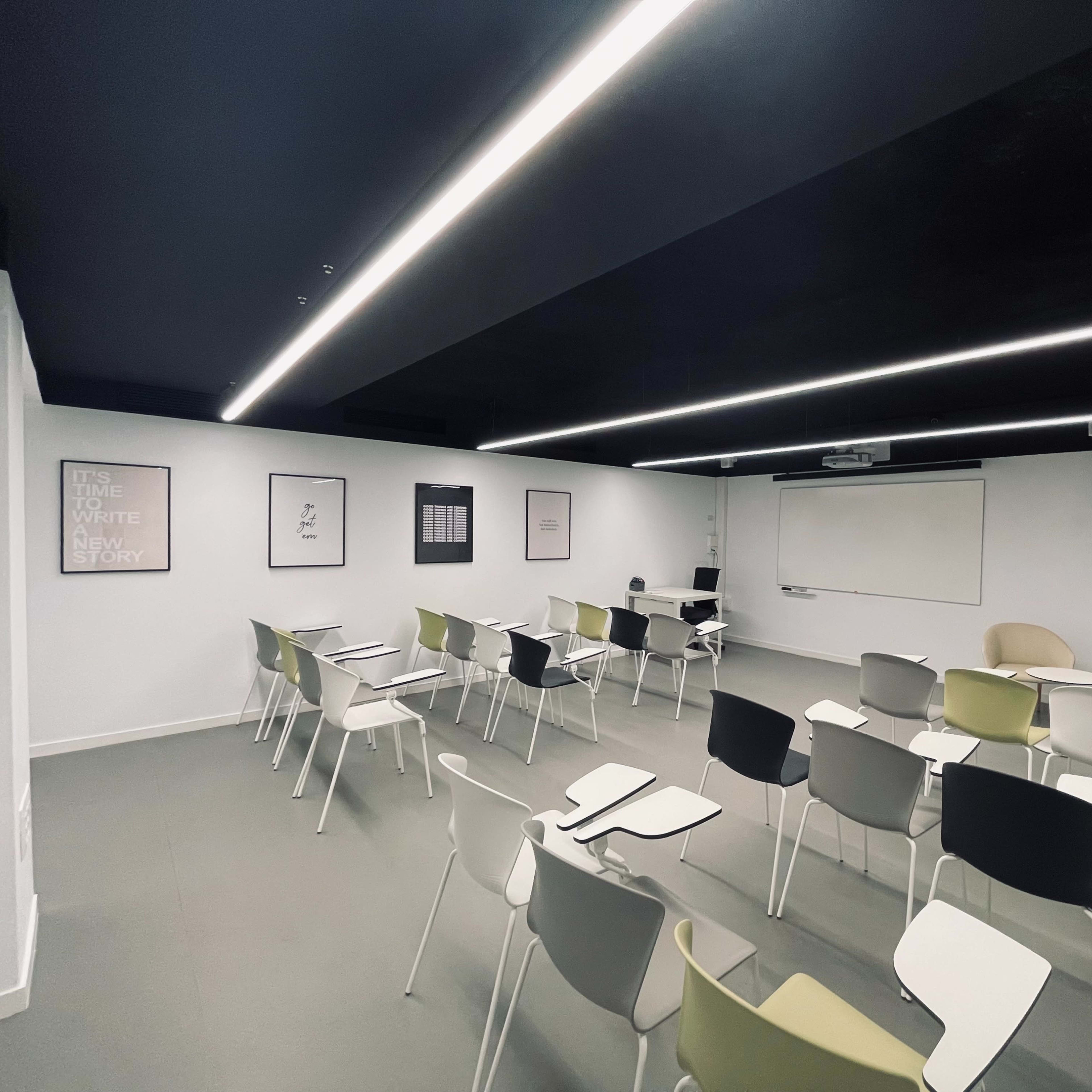 A modern classroom is arranged with rows of chairs and a whiteboard at the front, illuminated by linear ceiling lights.