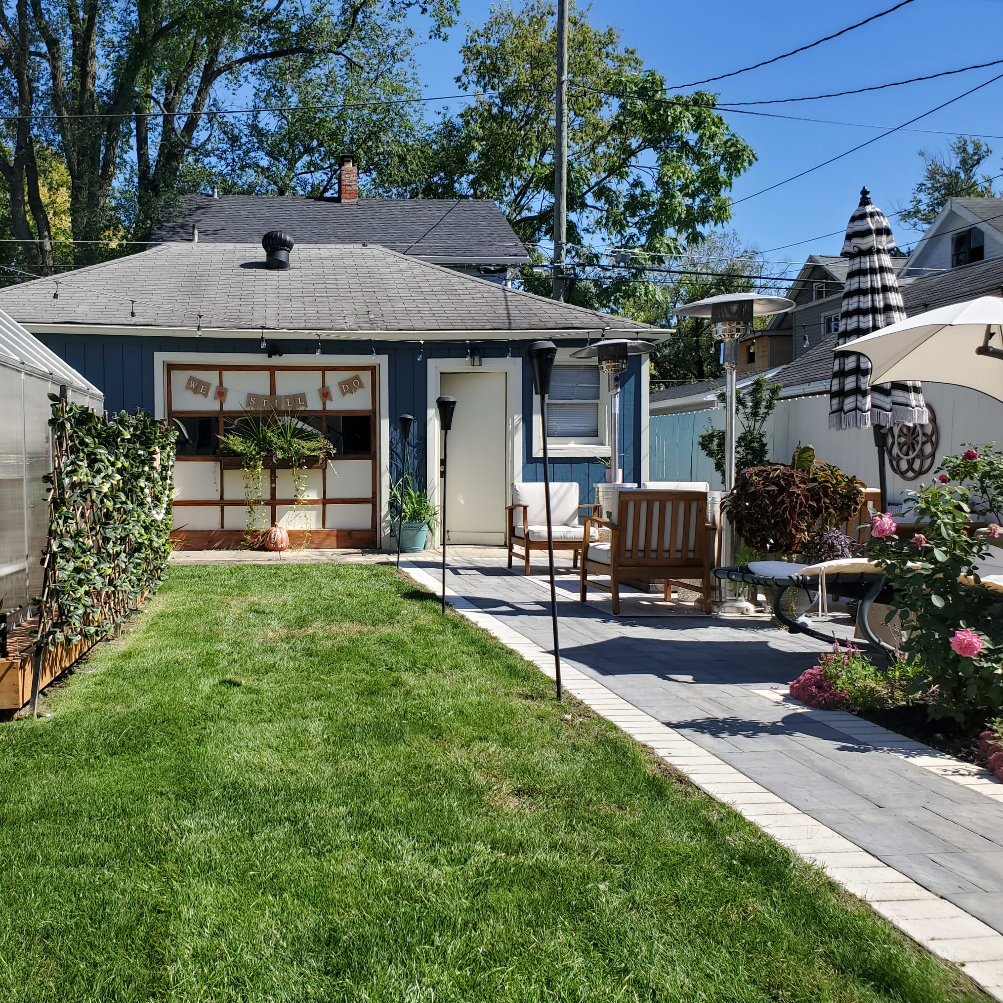 A landscaped backyard with a green lawn, a greenhouse on the left, and a patio area with outdoor furniture on the right.