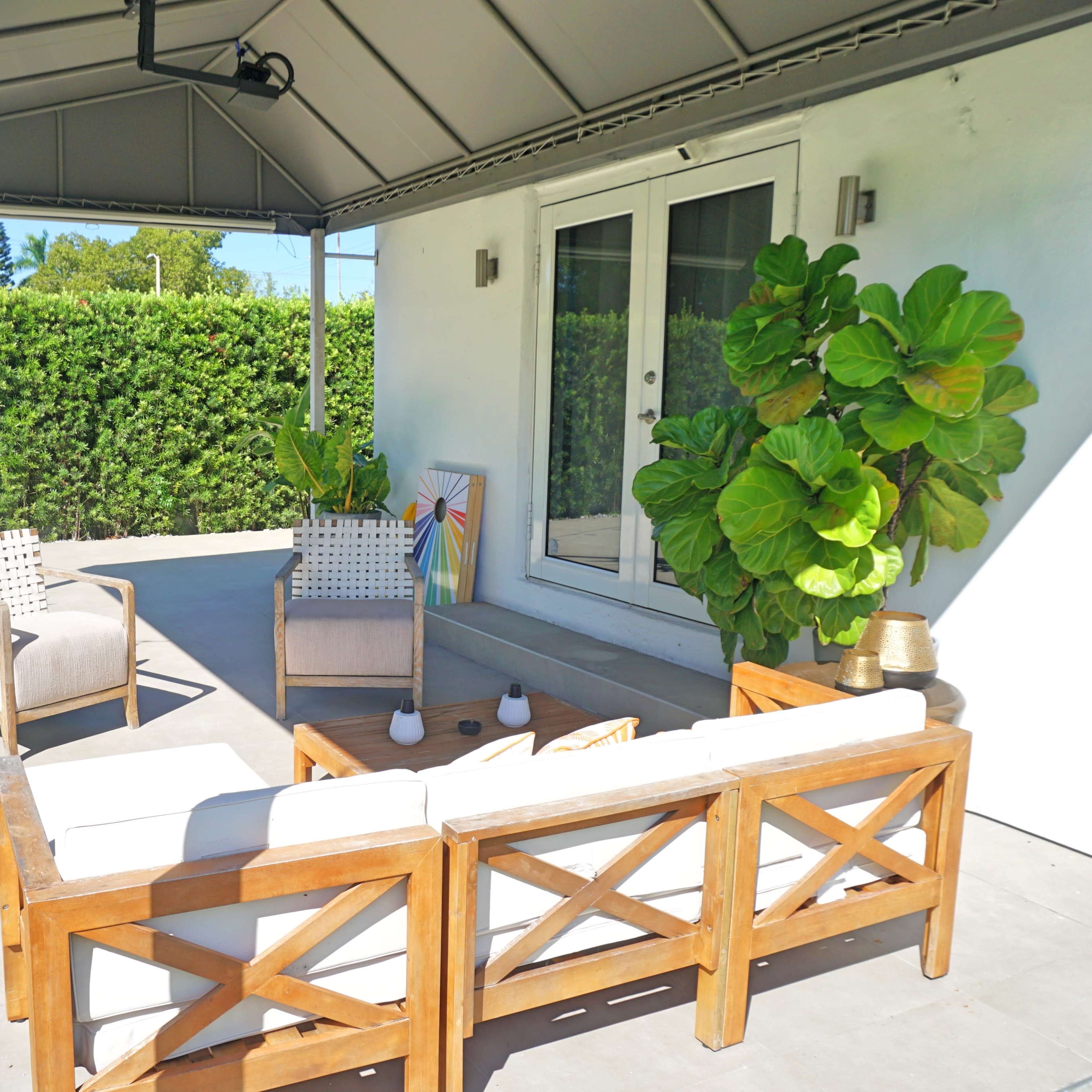 A covered patio features wooden furniture with cushions, a potted plant, and a doorway leading into a house.