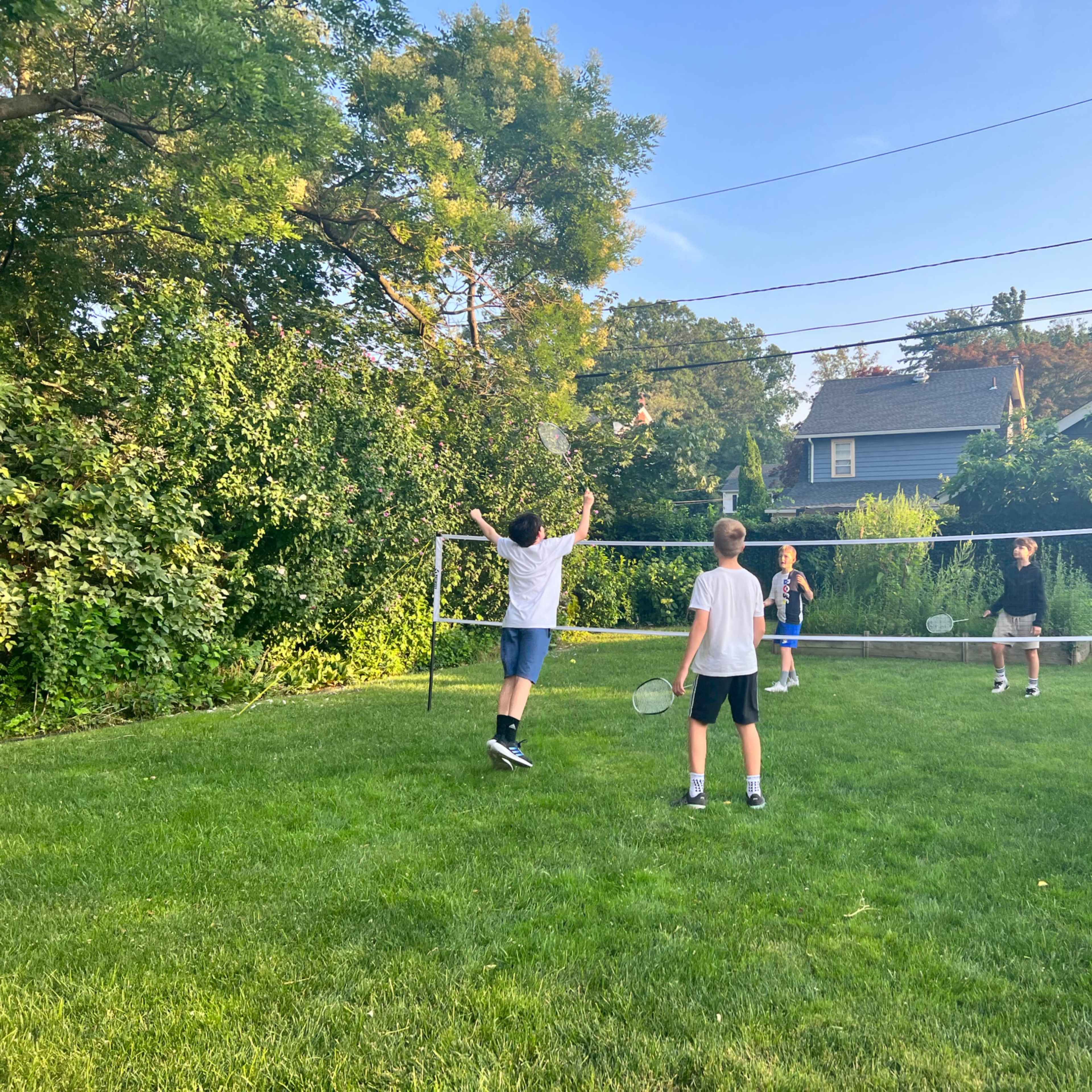 Four children are playing badminton on a grassy lawn surrounded by trees and houses.