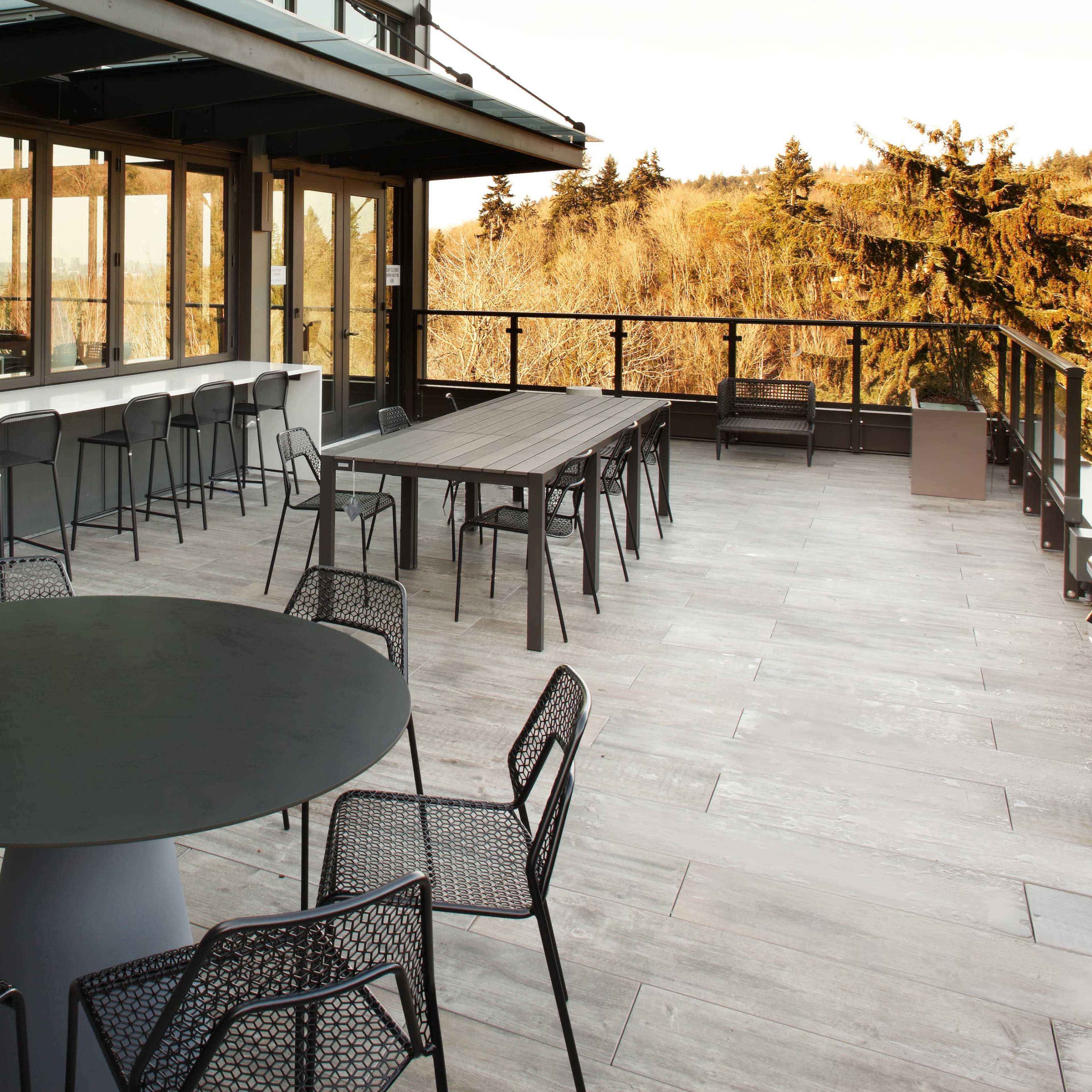 A modern outdoor terrace with a circular table and several chairs, along with a long rectangular table and additional seating, set against a backdrop of trees and a clear sky.