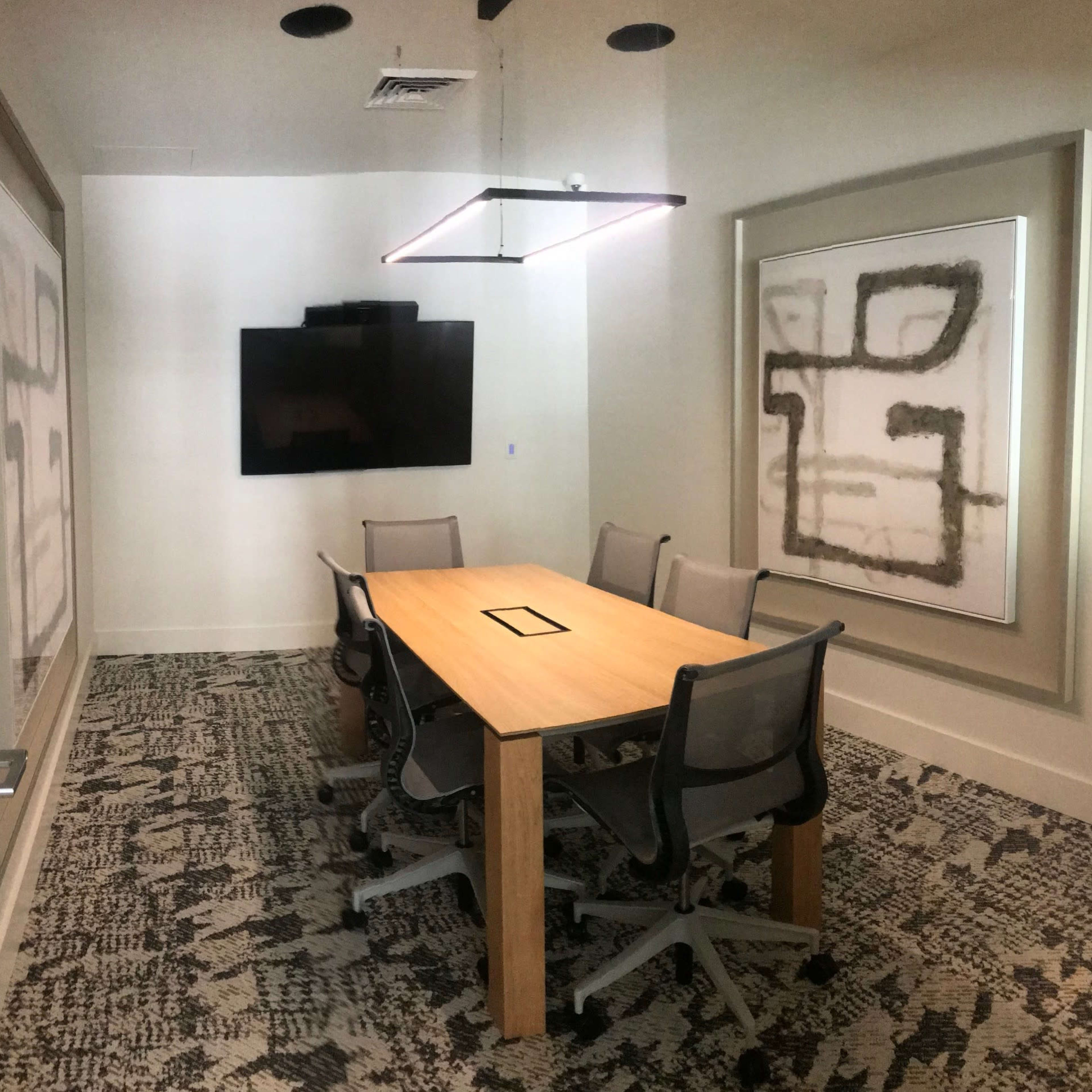 A modern conference room with a wooden table surrounded by four chairs, a large wall-mounted screen, and abstract art on the wall.