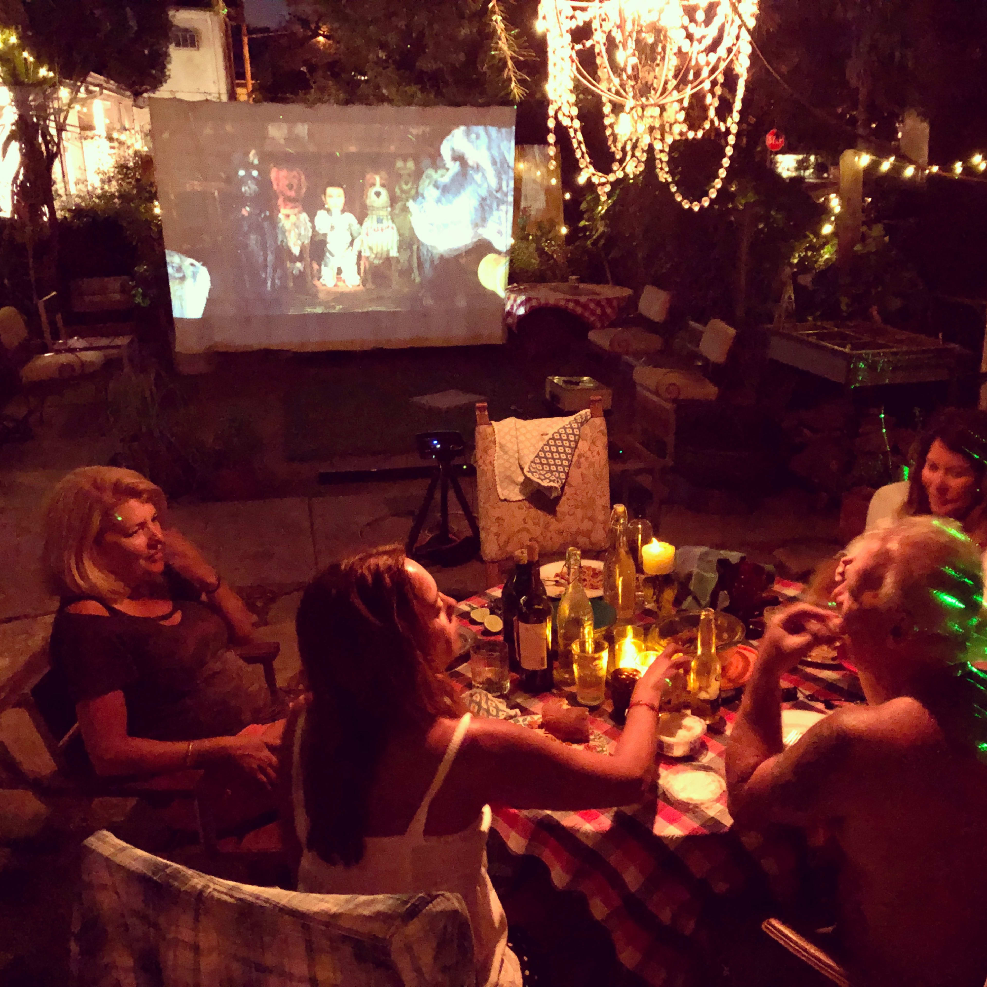 A group of five people sits around a table in a dimly lit outdoor setting, enjoying food and drinks while a movie is projected on a nearby wall.