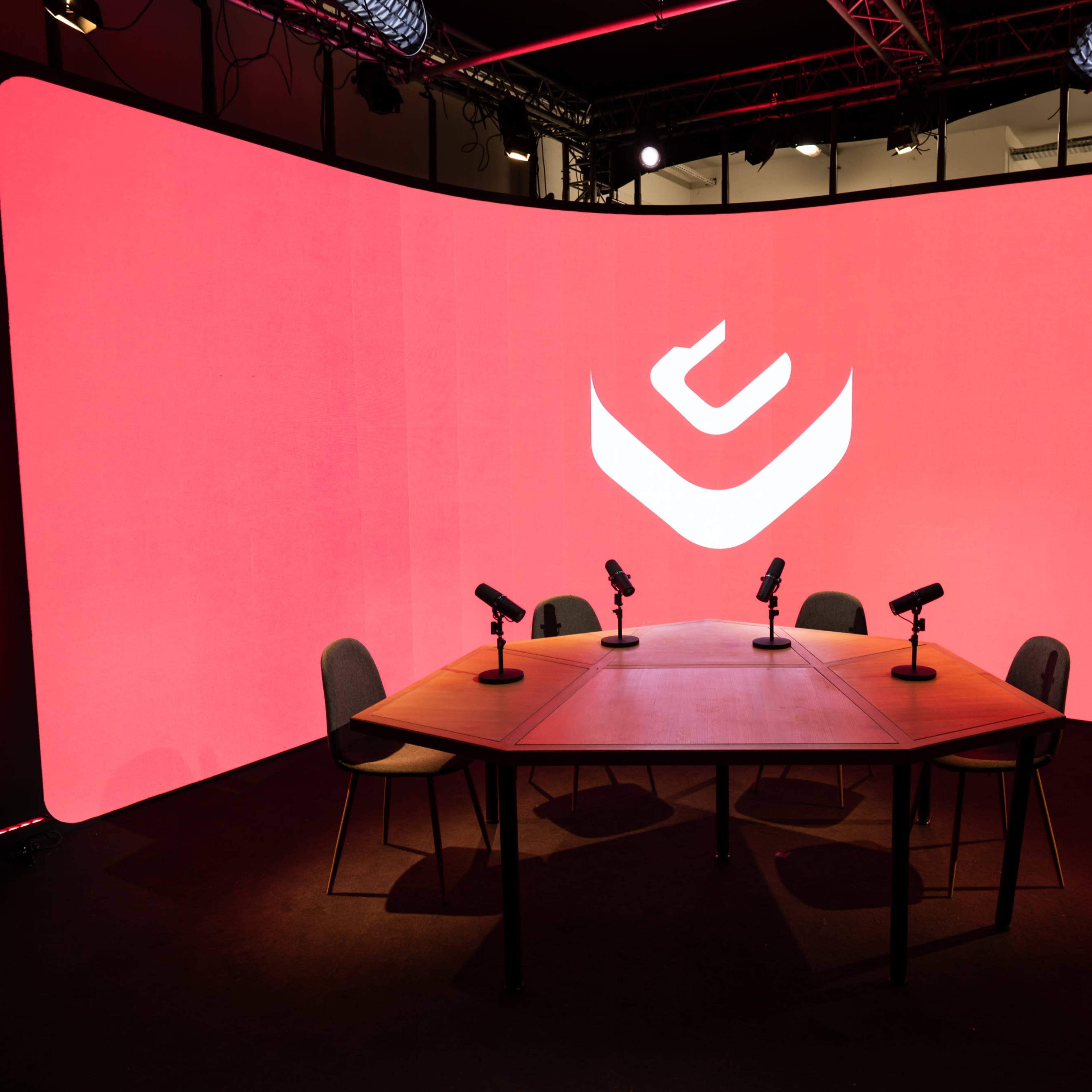 A circular wooden table with four chairs and microphones is set in front of a large red backdrop featuring a white logo.