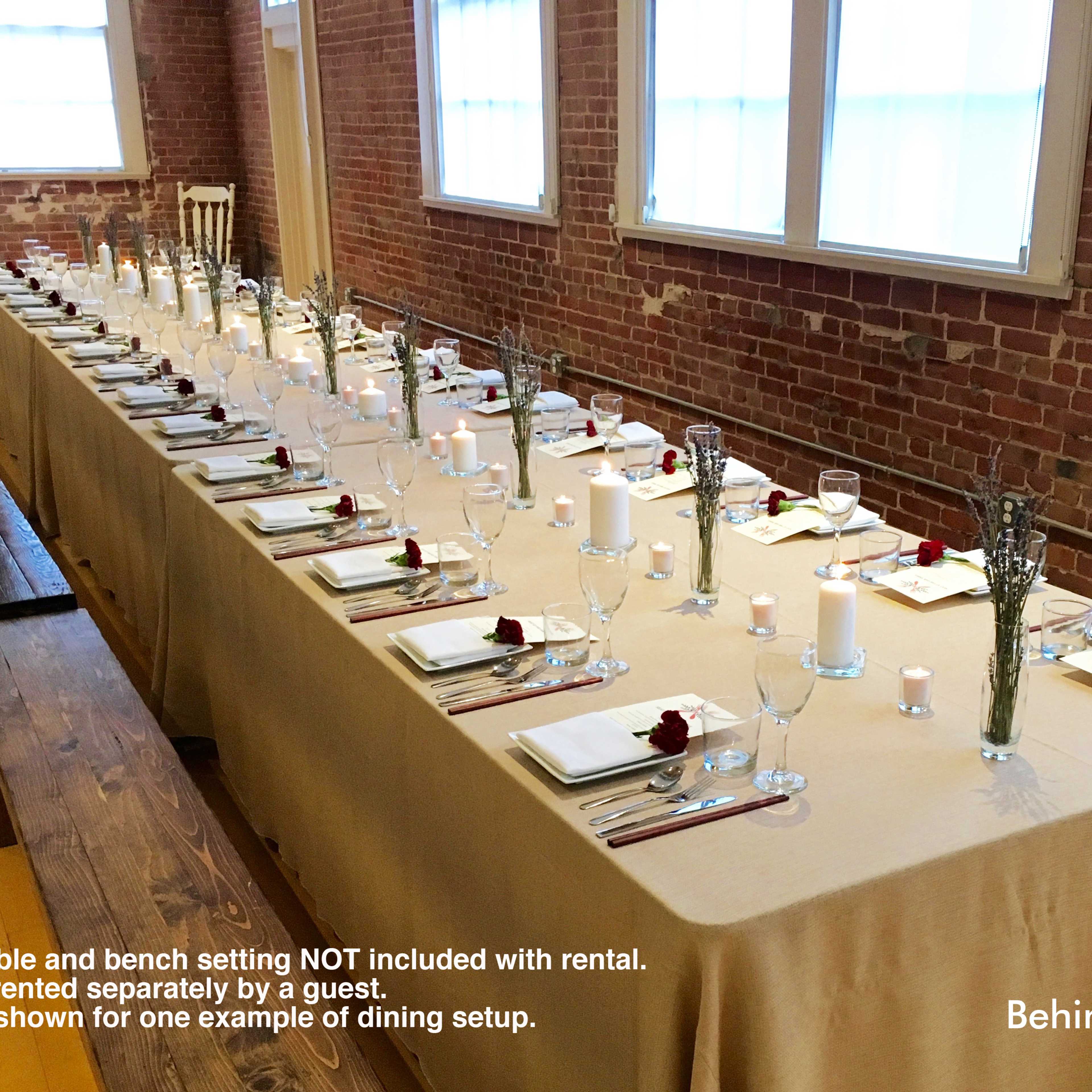 A long dining table is set with glasses, plates, and candles, arranged along a brick wall in a well-lit room.
