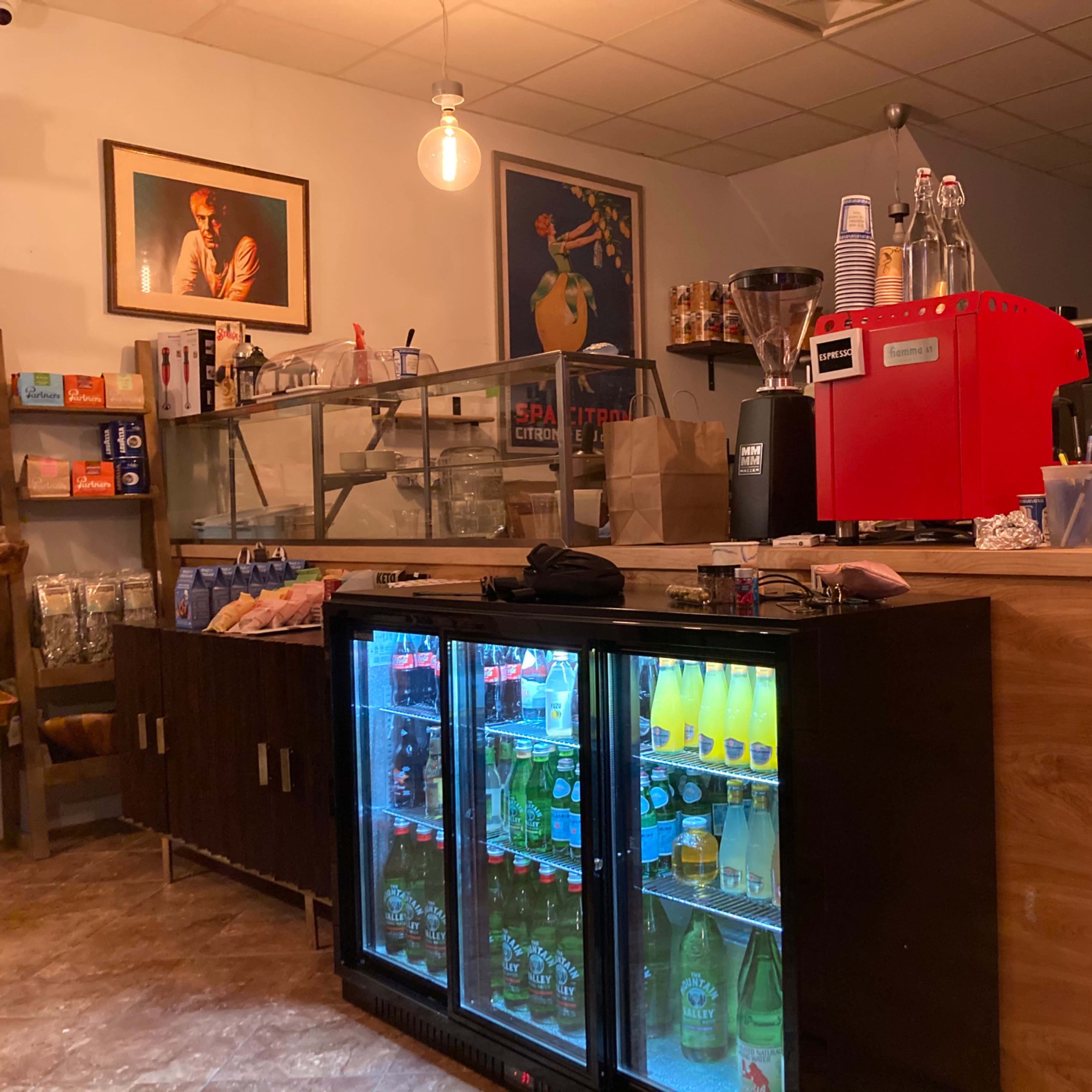 A small café interior featuring a wooden counter with a red espresso machine, glass display cases, and refrigerators containing beverages.