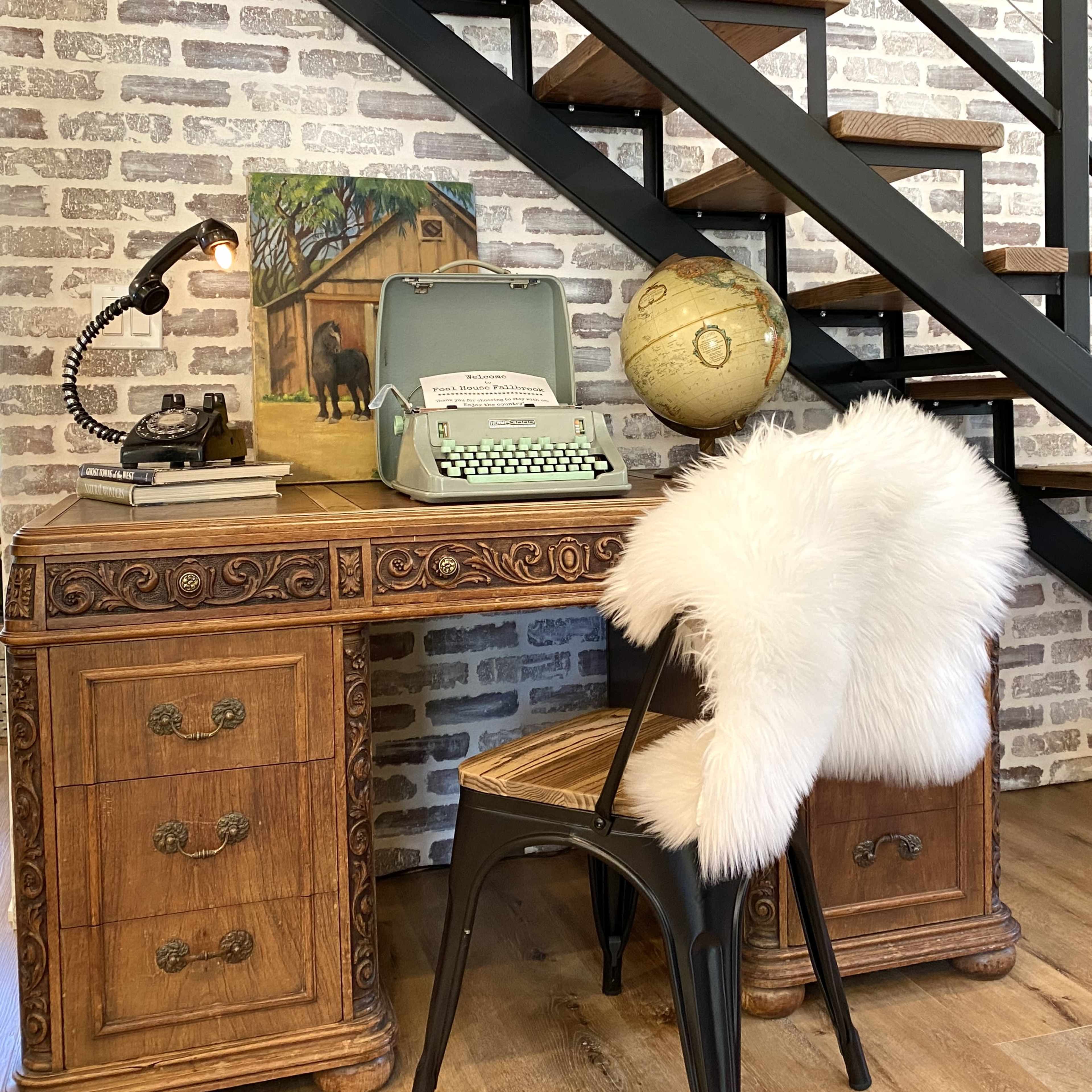 A vintage wooden desk with a typewriter and a globe sits beside a staircase with a brick wall background.