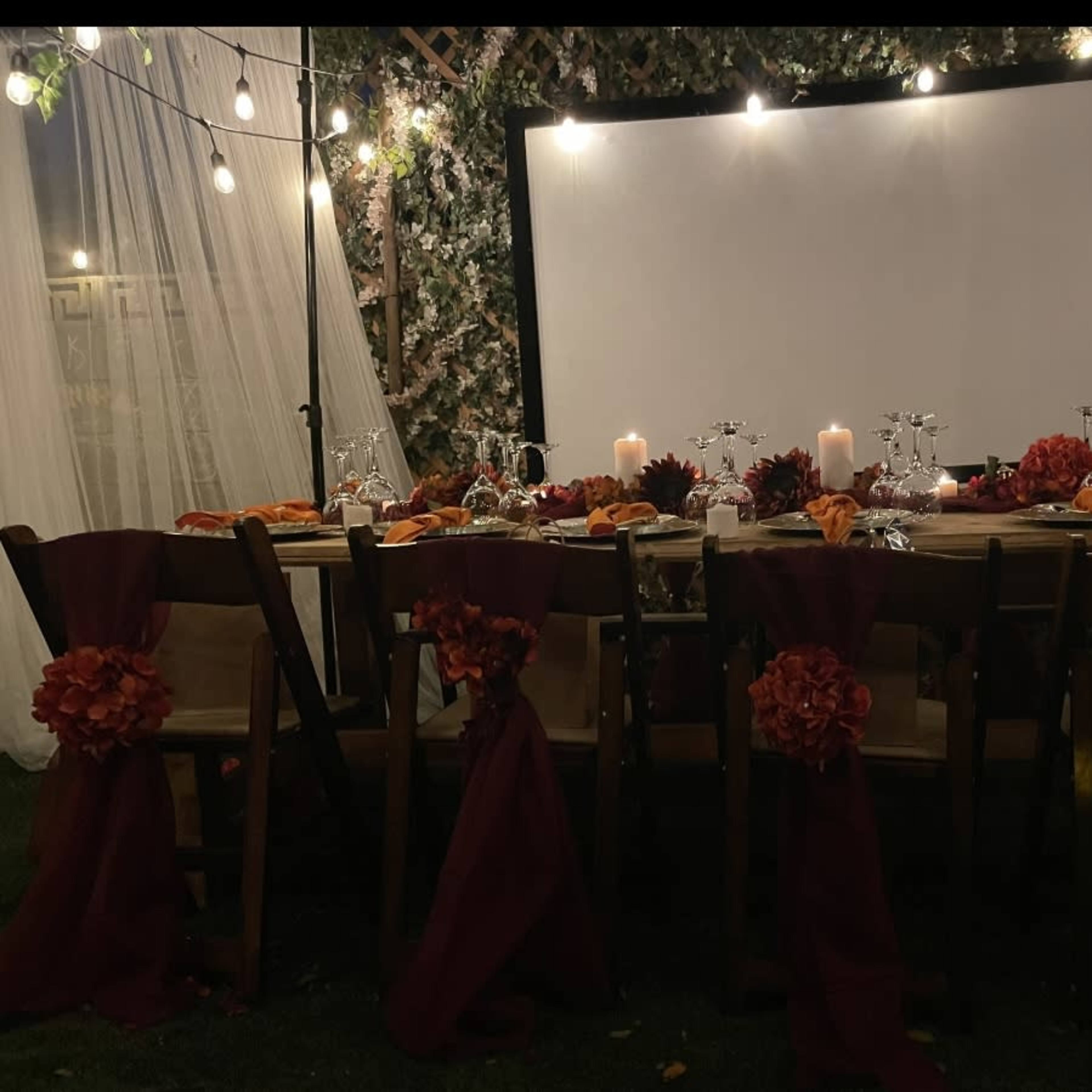 A long dining table is set up outdoors with candles, decorative flowers, and a large screen in the background, surrounded by draped fabric and string lights.