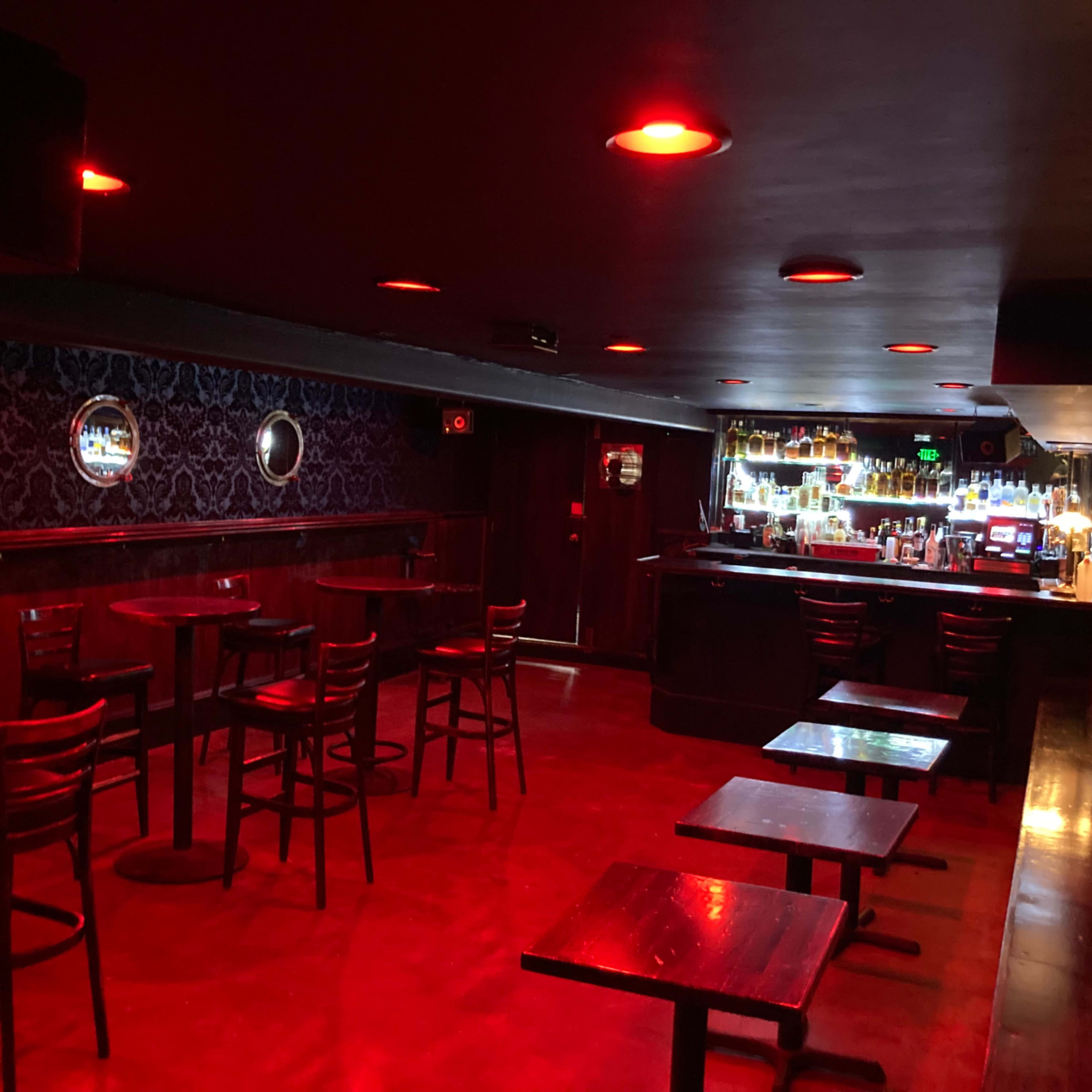 A dimly lit bar features a row of empty tables and a lighted bar counter in the background.