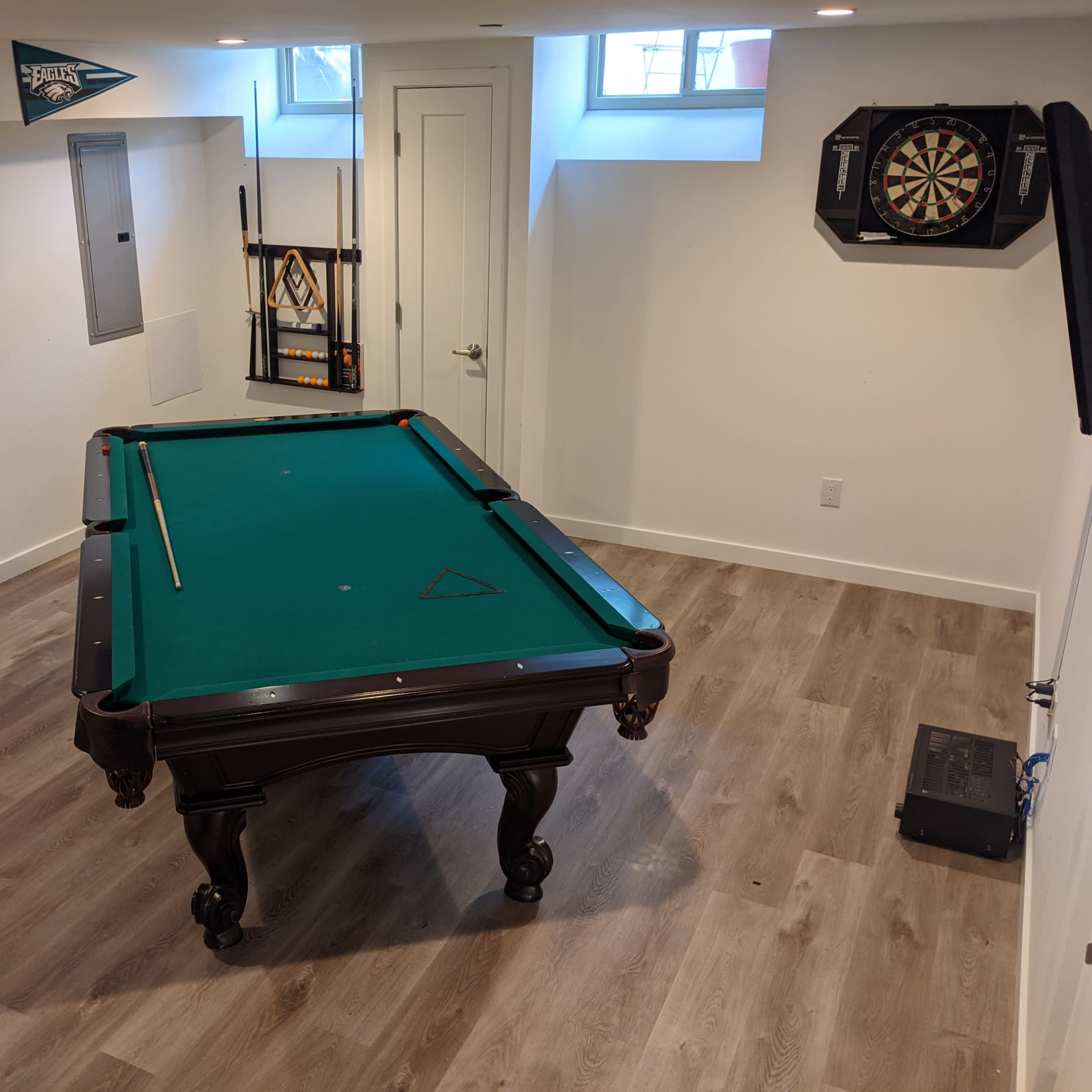 A well-lit basement with a green felt pool table, a dartboard on the wall, and bar stools nearby.