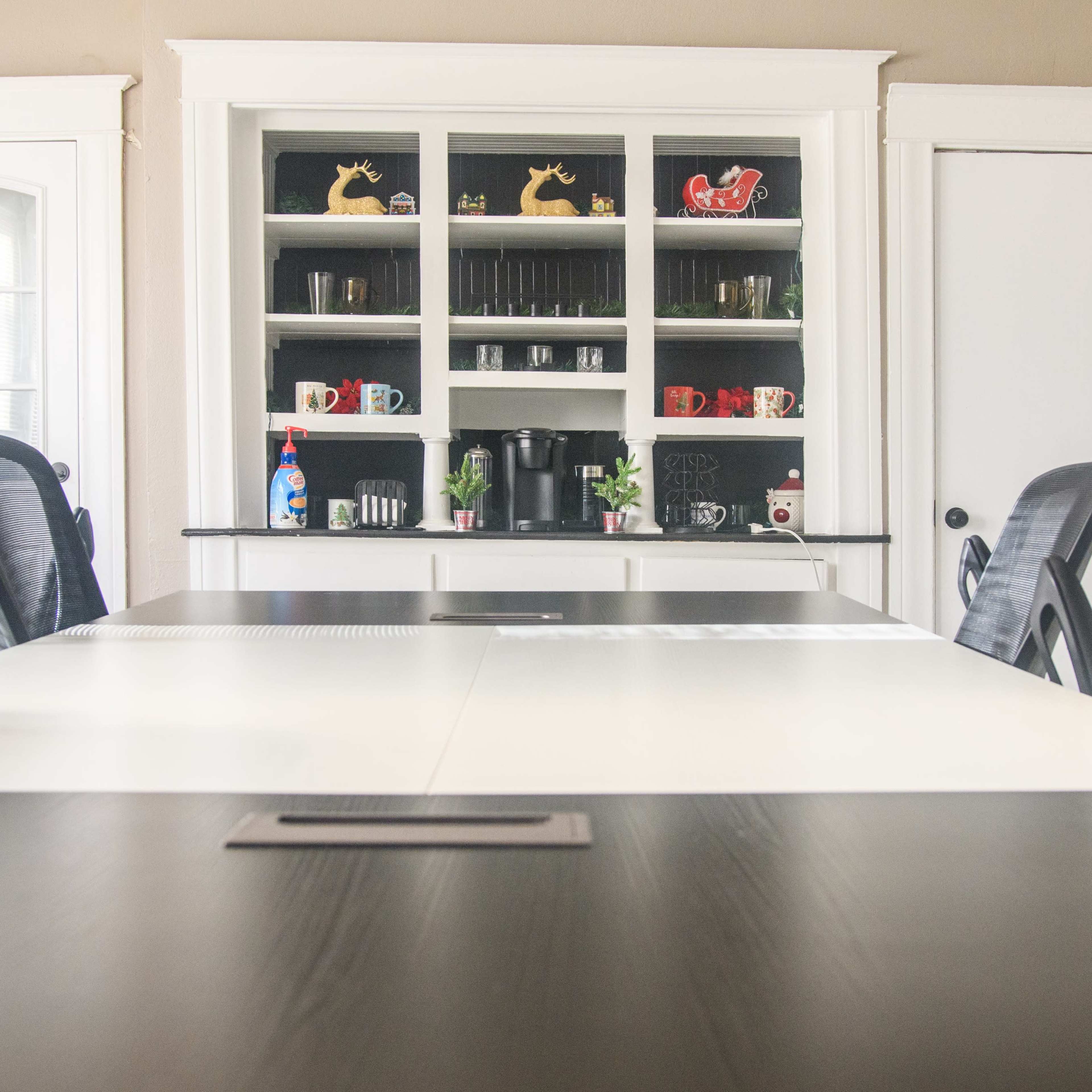 A modern conference room with a long table, black chairs, and a shelving unit displaying mugs and other items in the background.