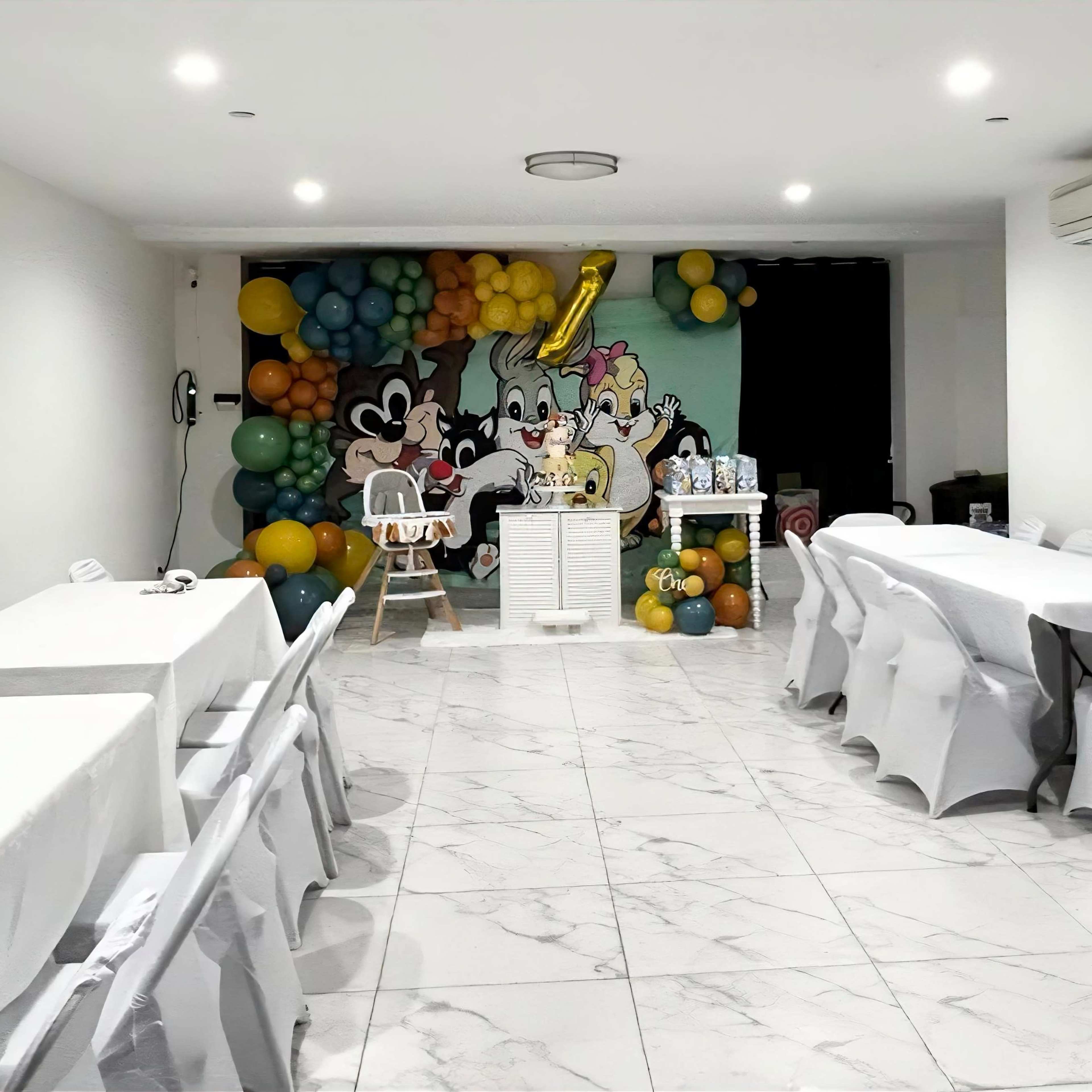 A decorated room with white tables and chairs set up, featuring a colorful balloon wall and a backdrop with cartoon characters.
