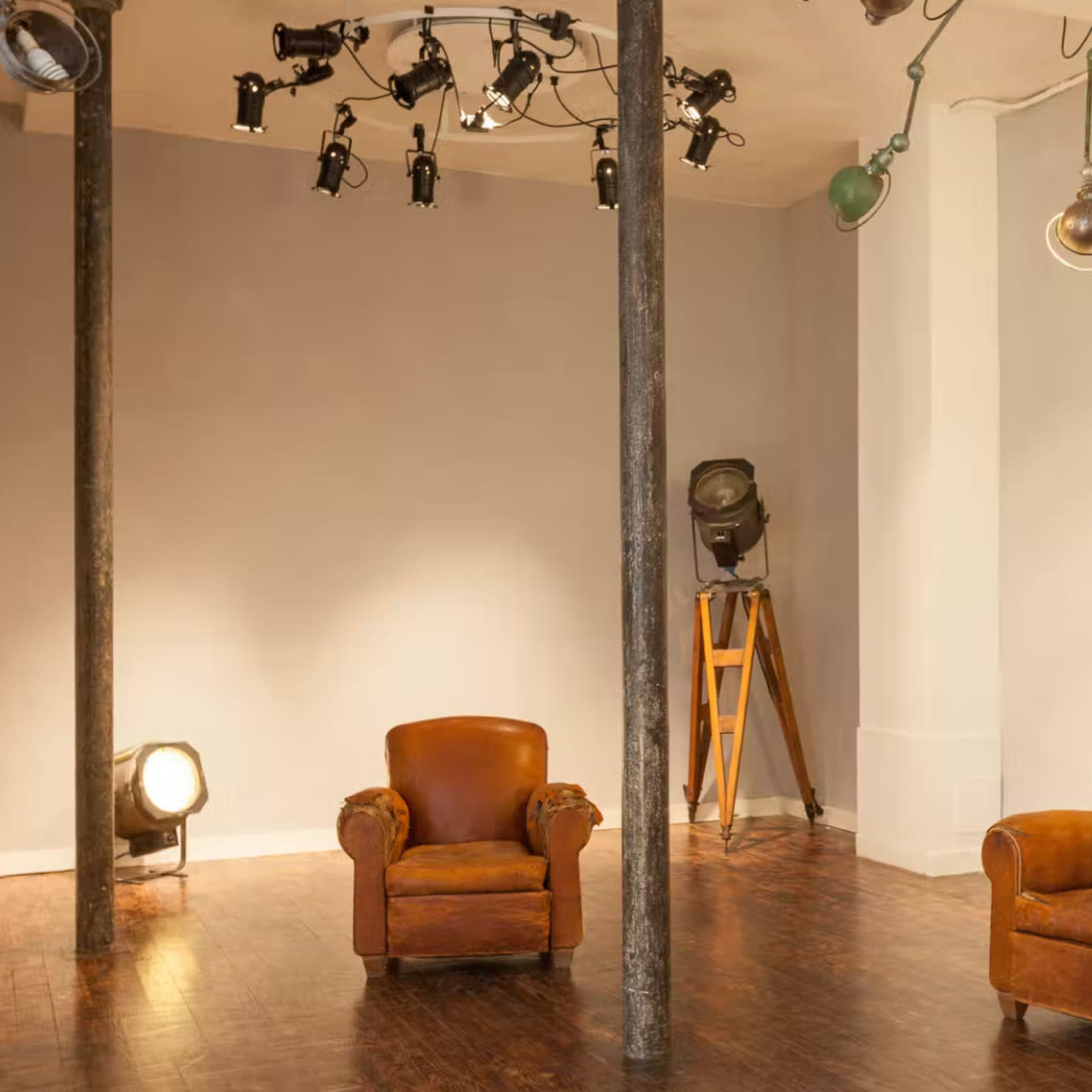A minimalist interior space featuring two brown leather armchairs, a tall wooden tripod light stand, and various mounted lights on the ceiling.
