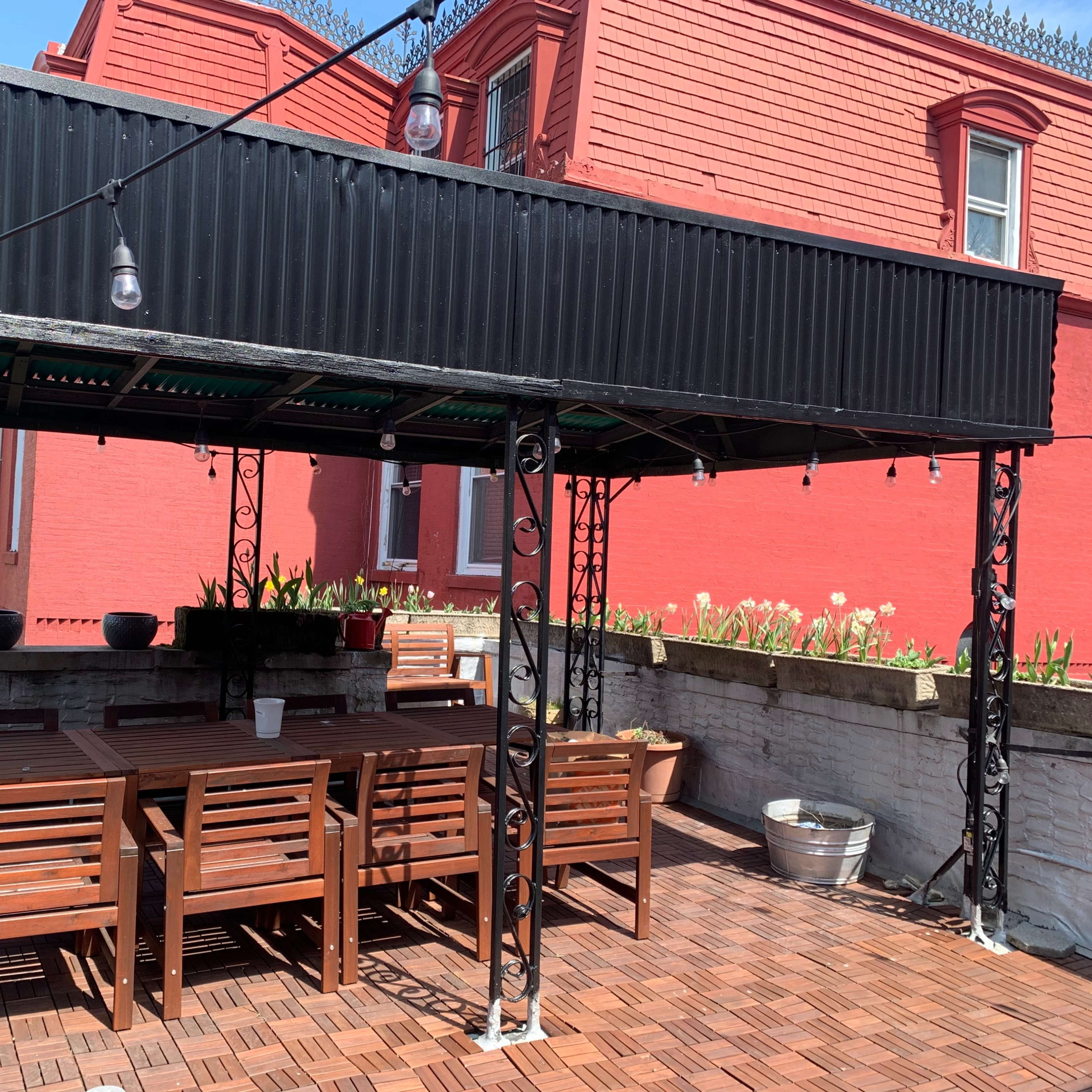 A wooden outdoor seating area with a covered gazebo and string lights, surrounded by a garden with potted plants against a red brick wall.