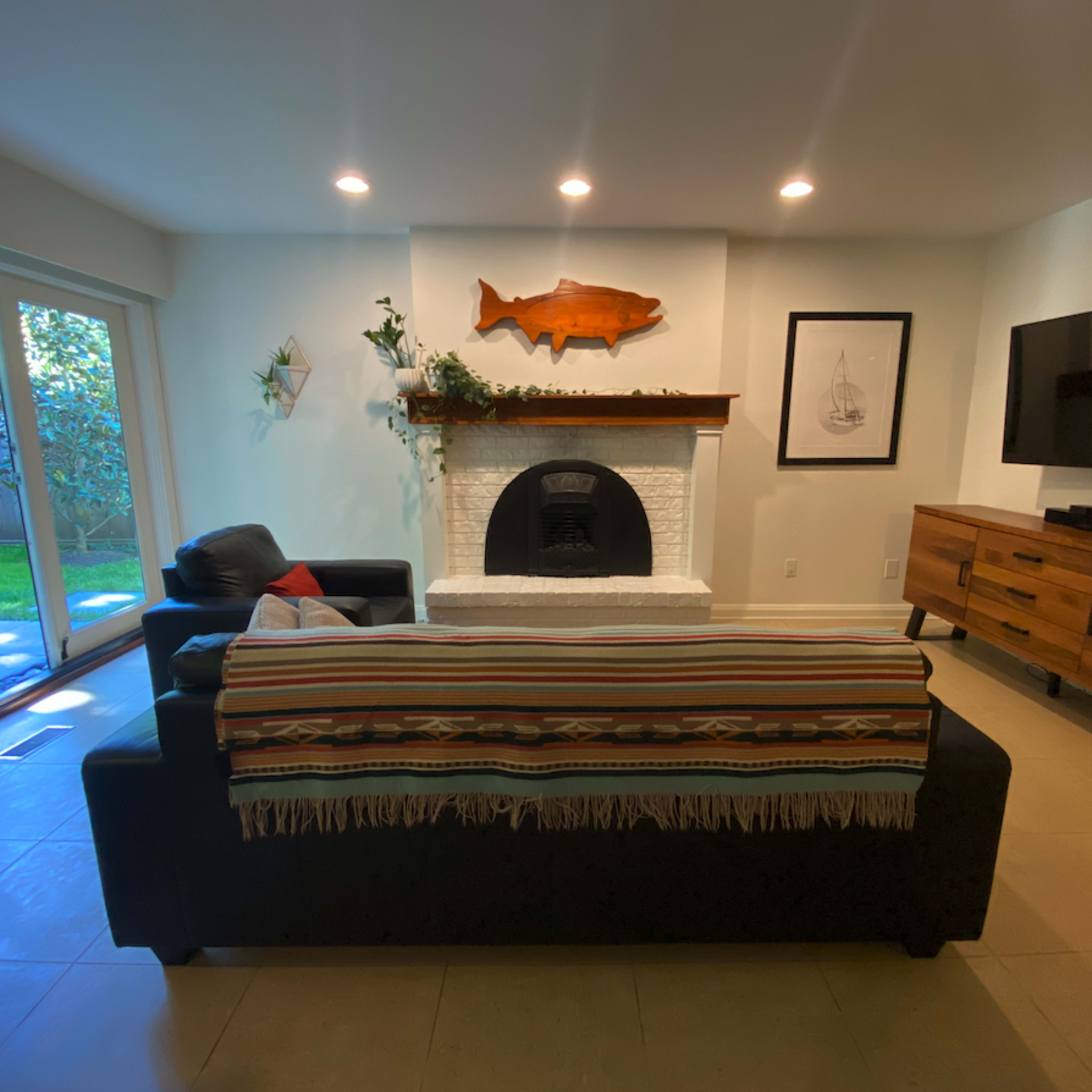 The living room features a black sofa with a striped blanket, a white brick fireplace with a wooden fish mounted above it, and a large TV mounted on the wall beside a wooden media console.