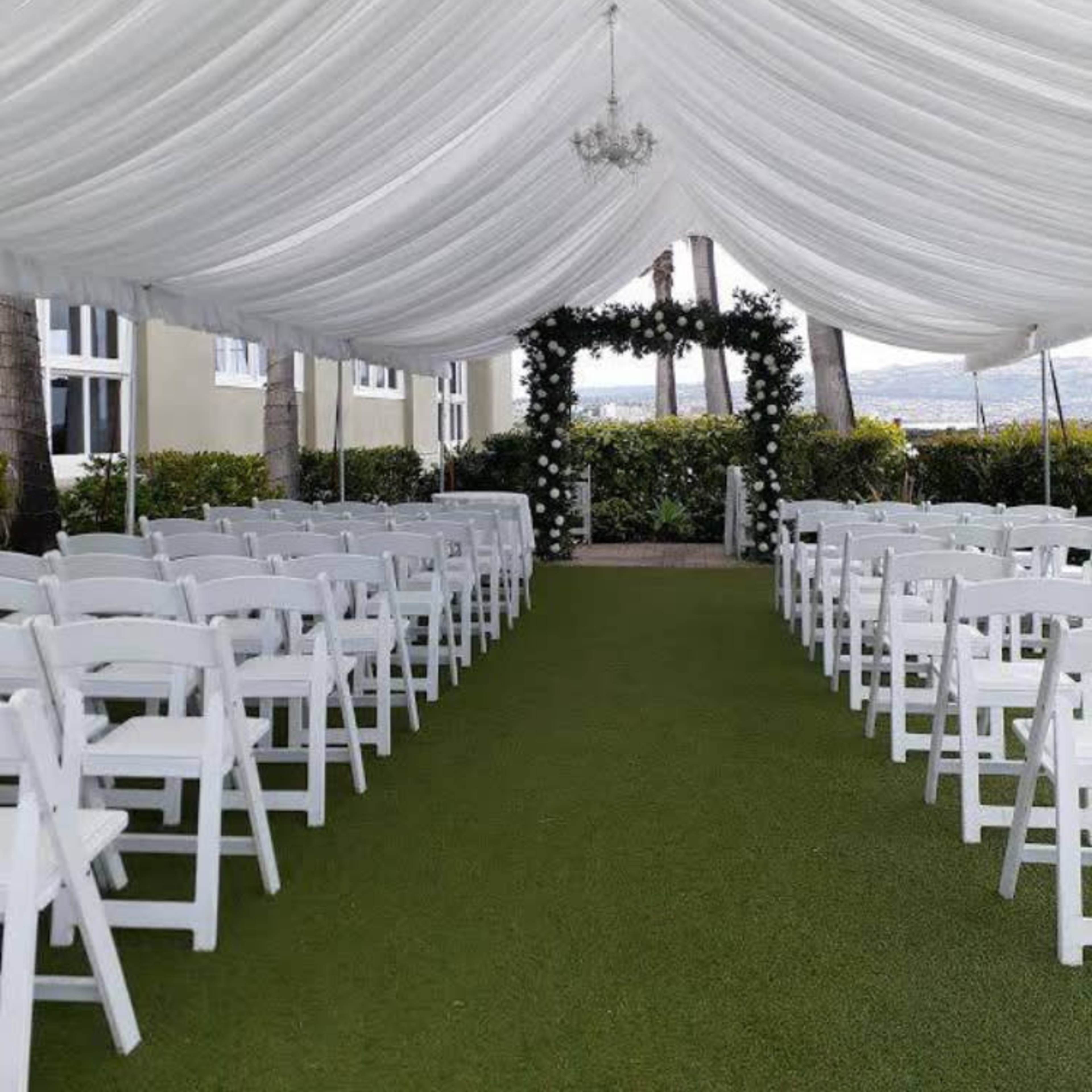 A tented outdoor venue features rows of white chairs set on green grass, leading to an arch adorned with greenery.