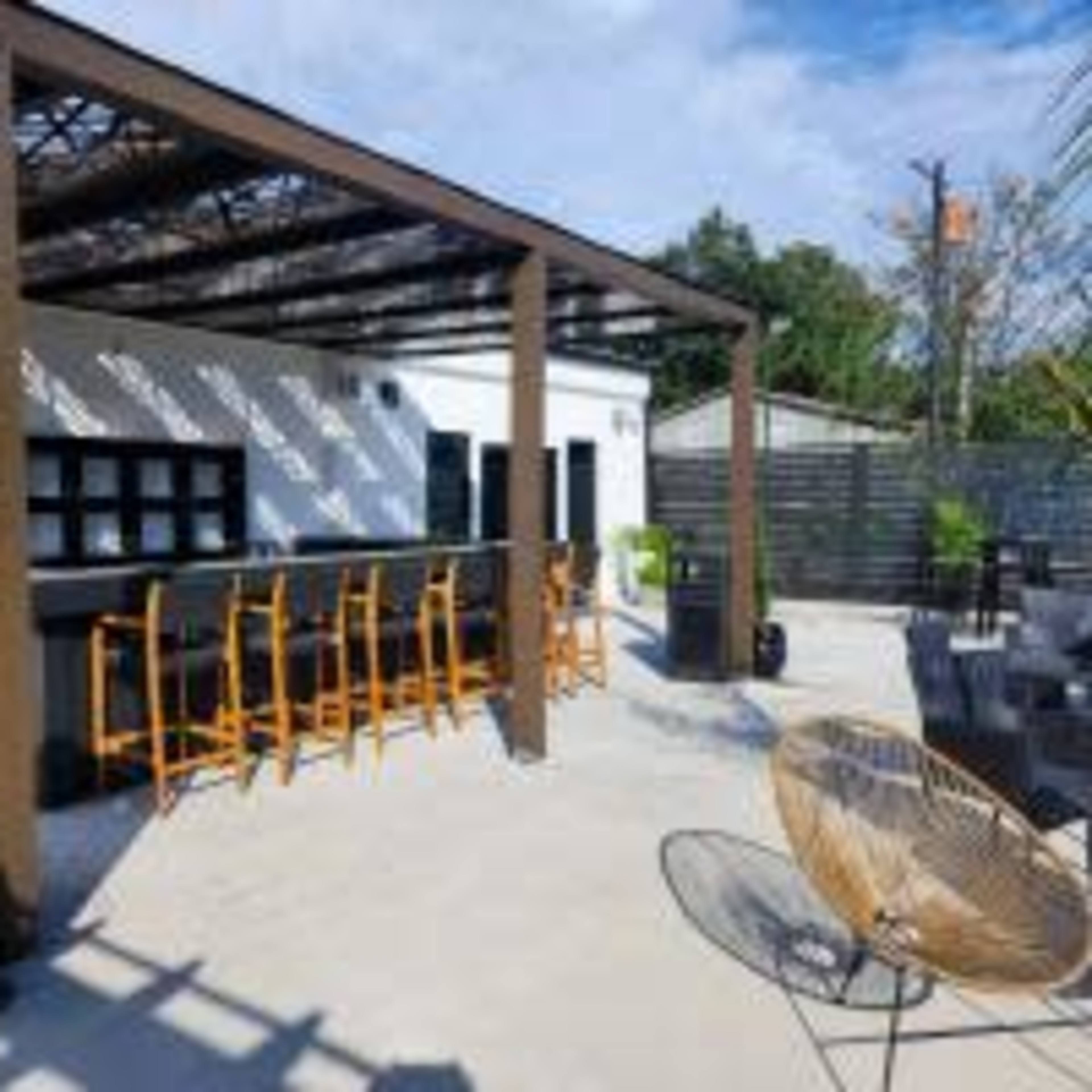 An outdoor bar area with wooden framing, high stools, and a stone countertop, surrounded by a landscaped patio.