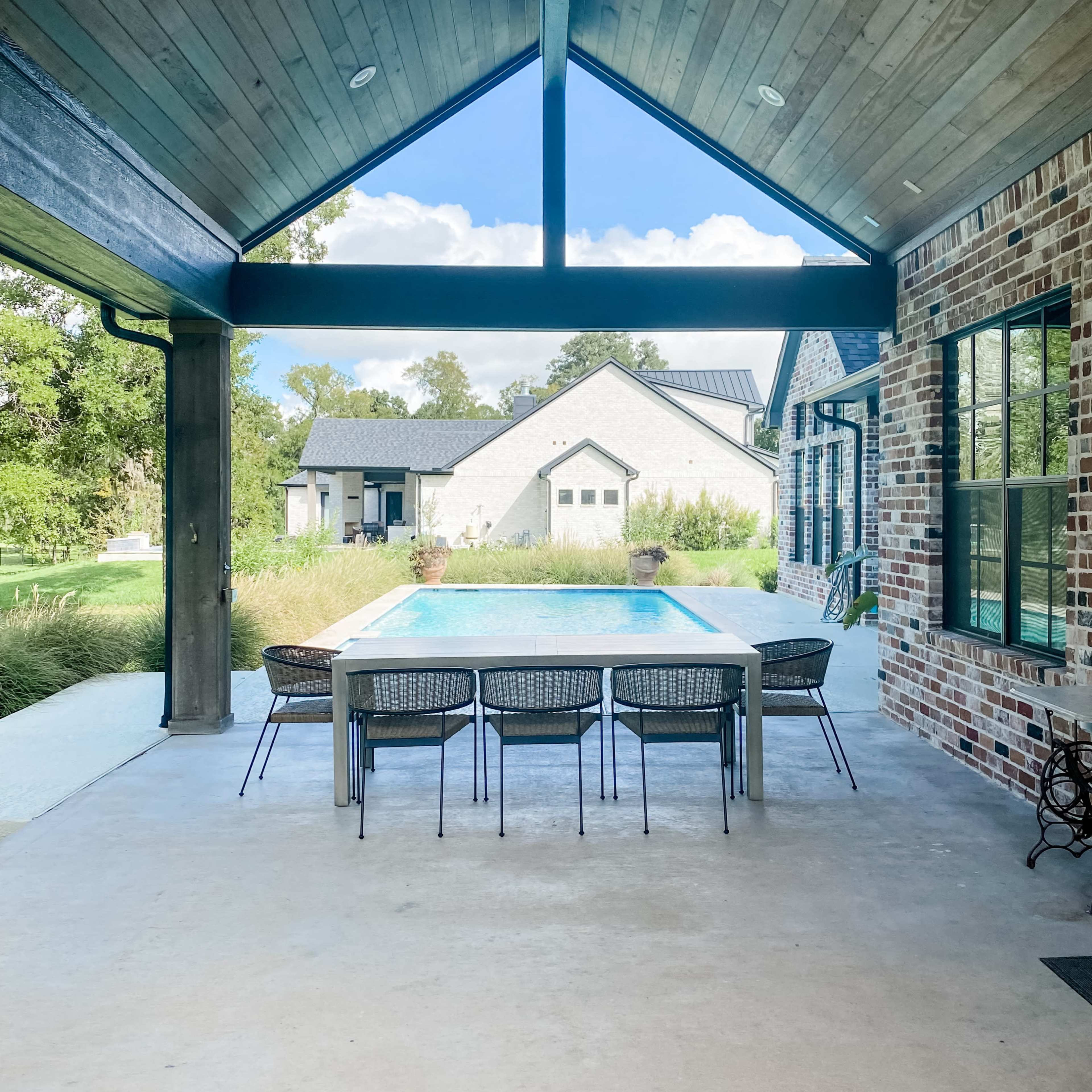 An outdoor patio area with a long table and chairs facing a swimming pool, framed by a high wooden ceiling and large windows.