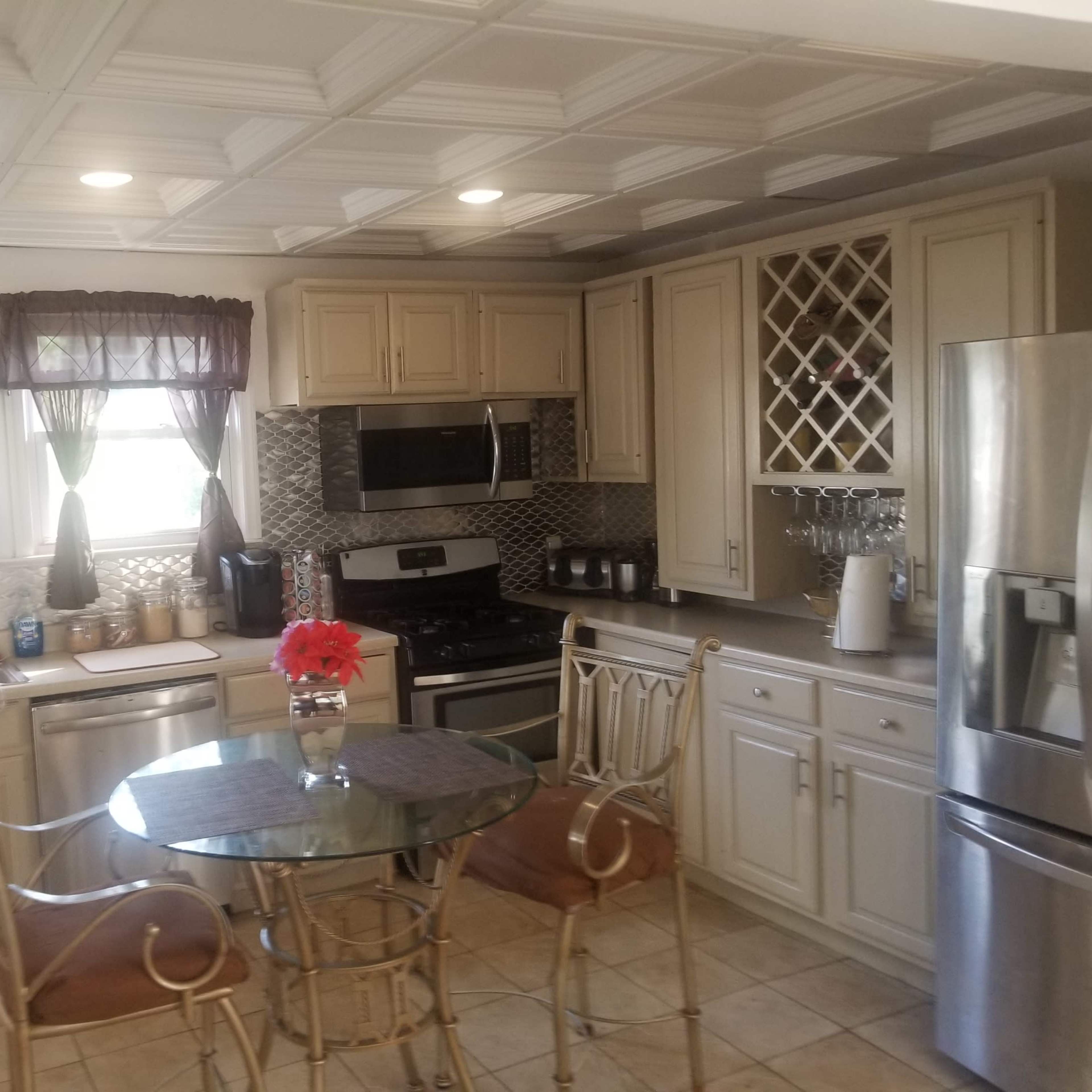 A modern kitchen featuring beige cabinetry, stainless steel appliances, a glass-top dining table, and window treatments with dark fabric.