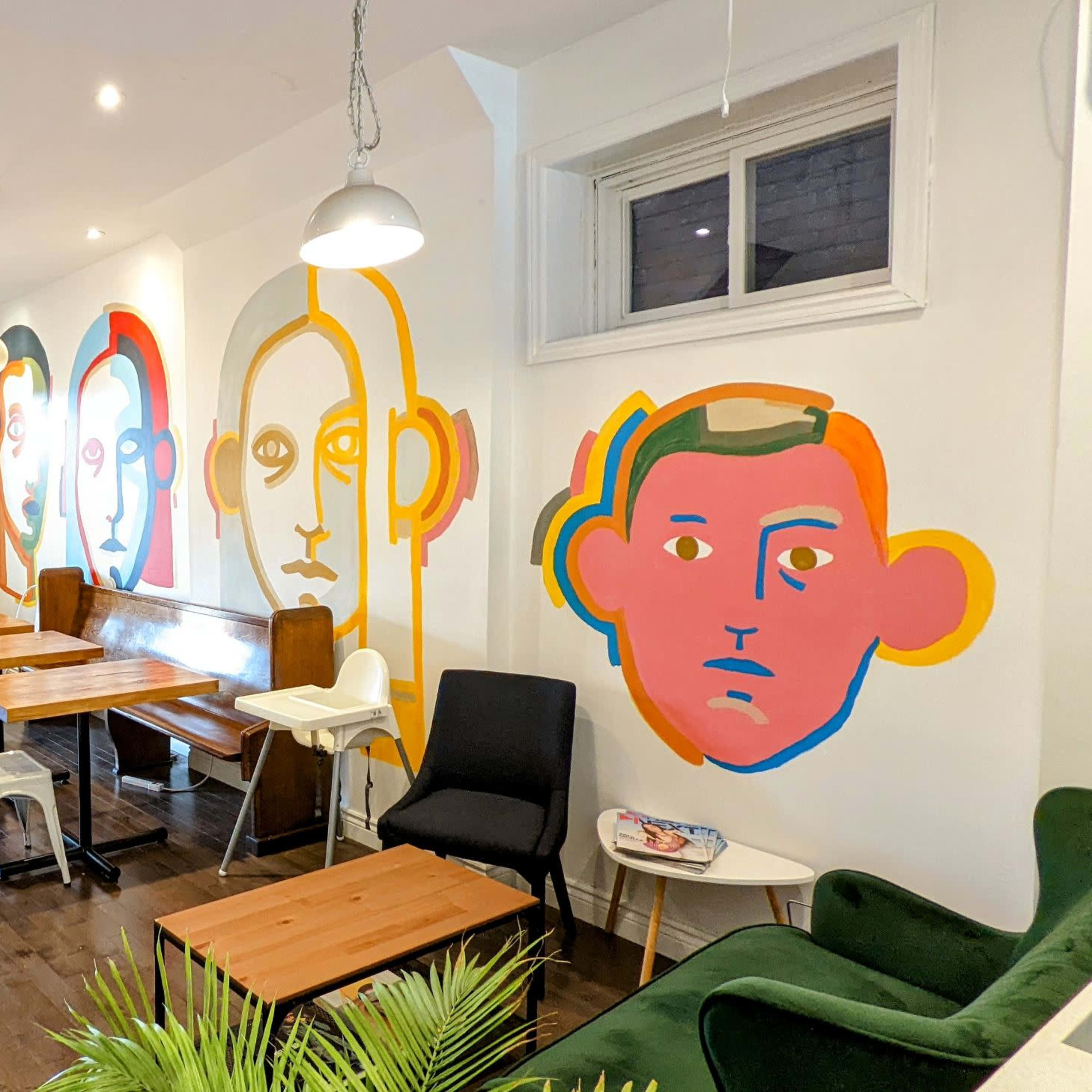A modern café interior featuring wooden tables, a staircase, and colorful mural artwork of stylized faces on the walls.