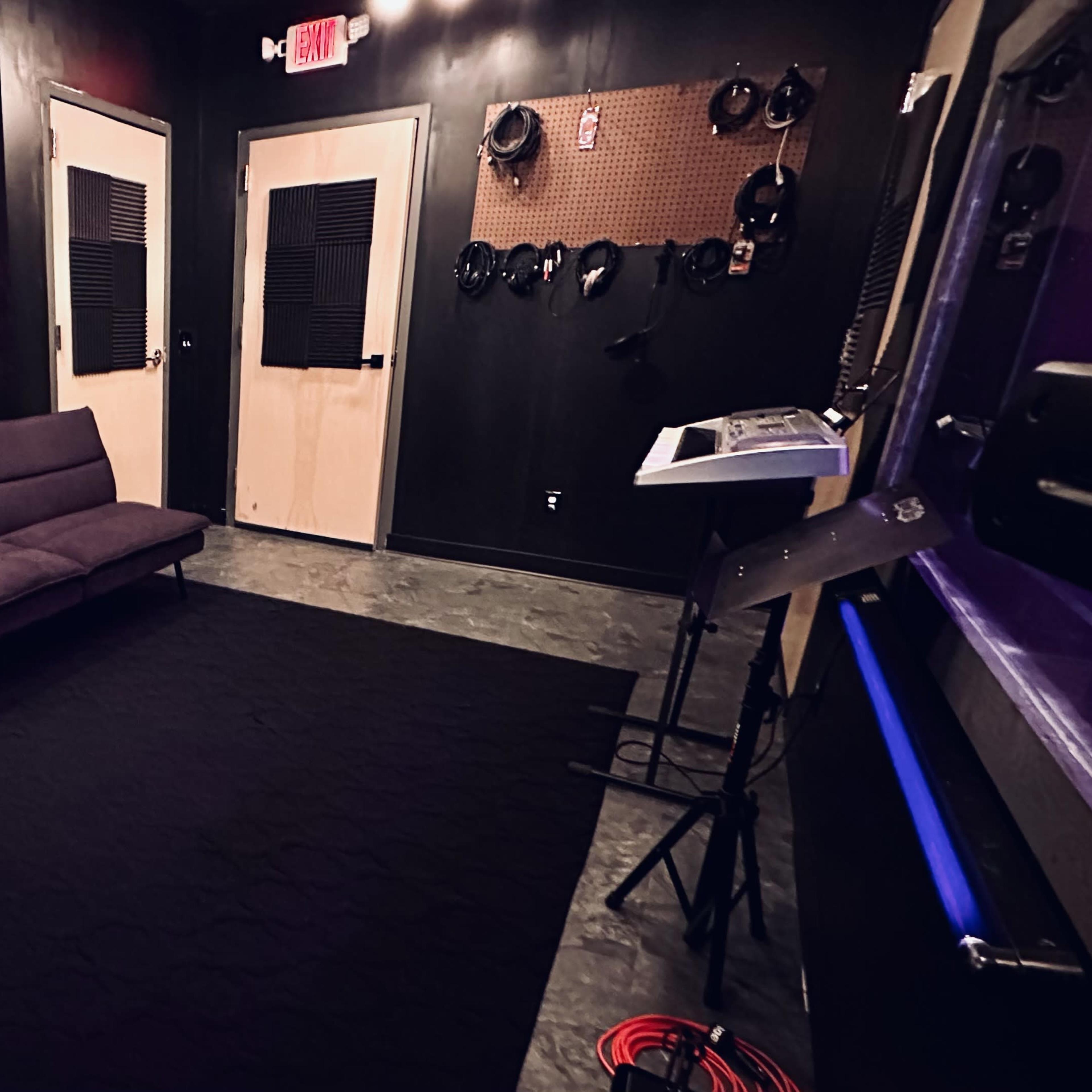 A small recording studio featuring black walls, a purple couch, soundproofing panels, and musical equipment arranged against the walls.