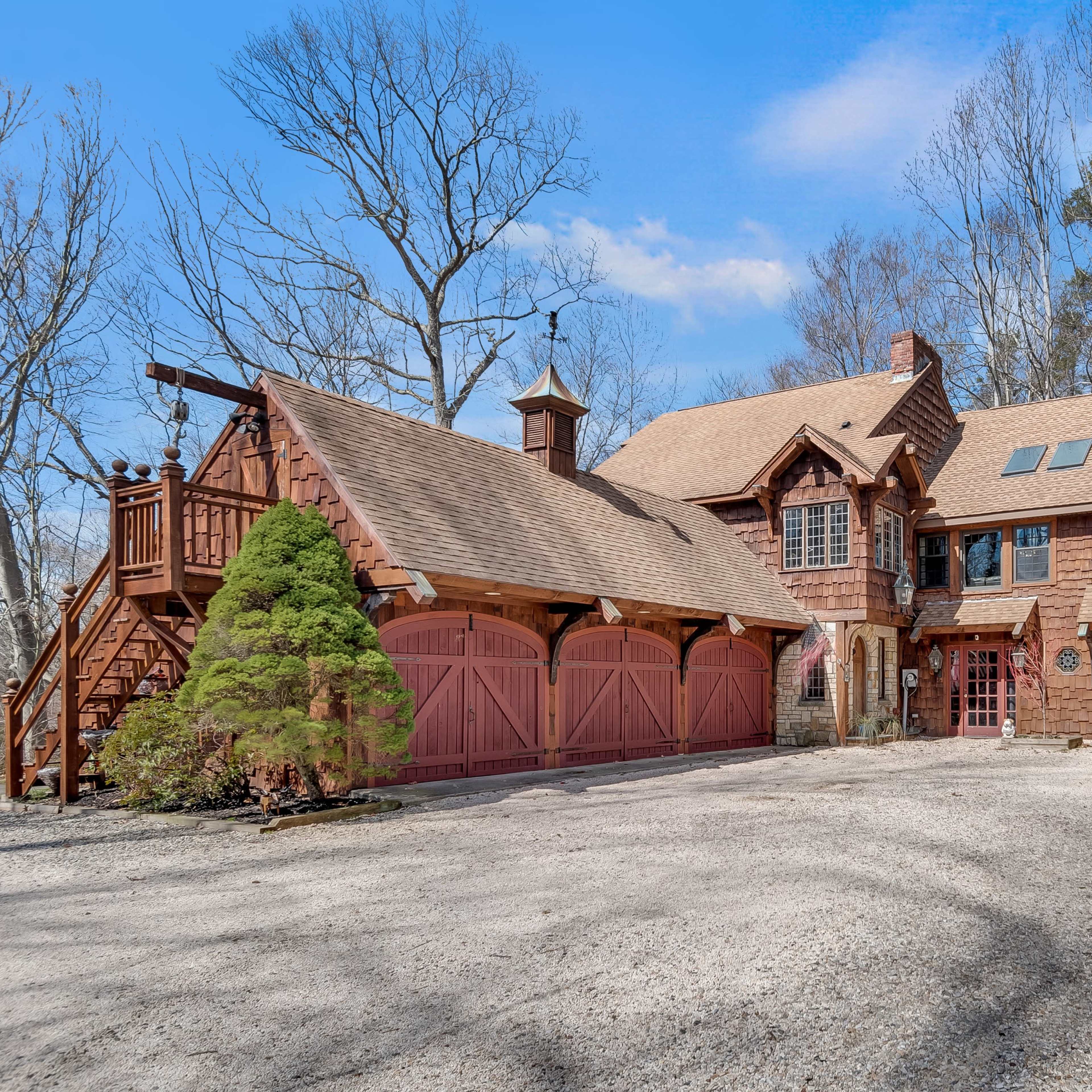 A large, rustic-style home with a gravel driveway and red garage doors is surrounded by bare trees.